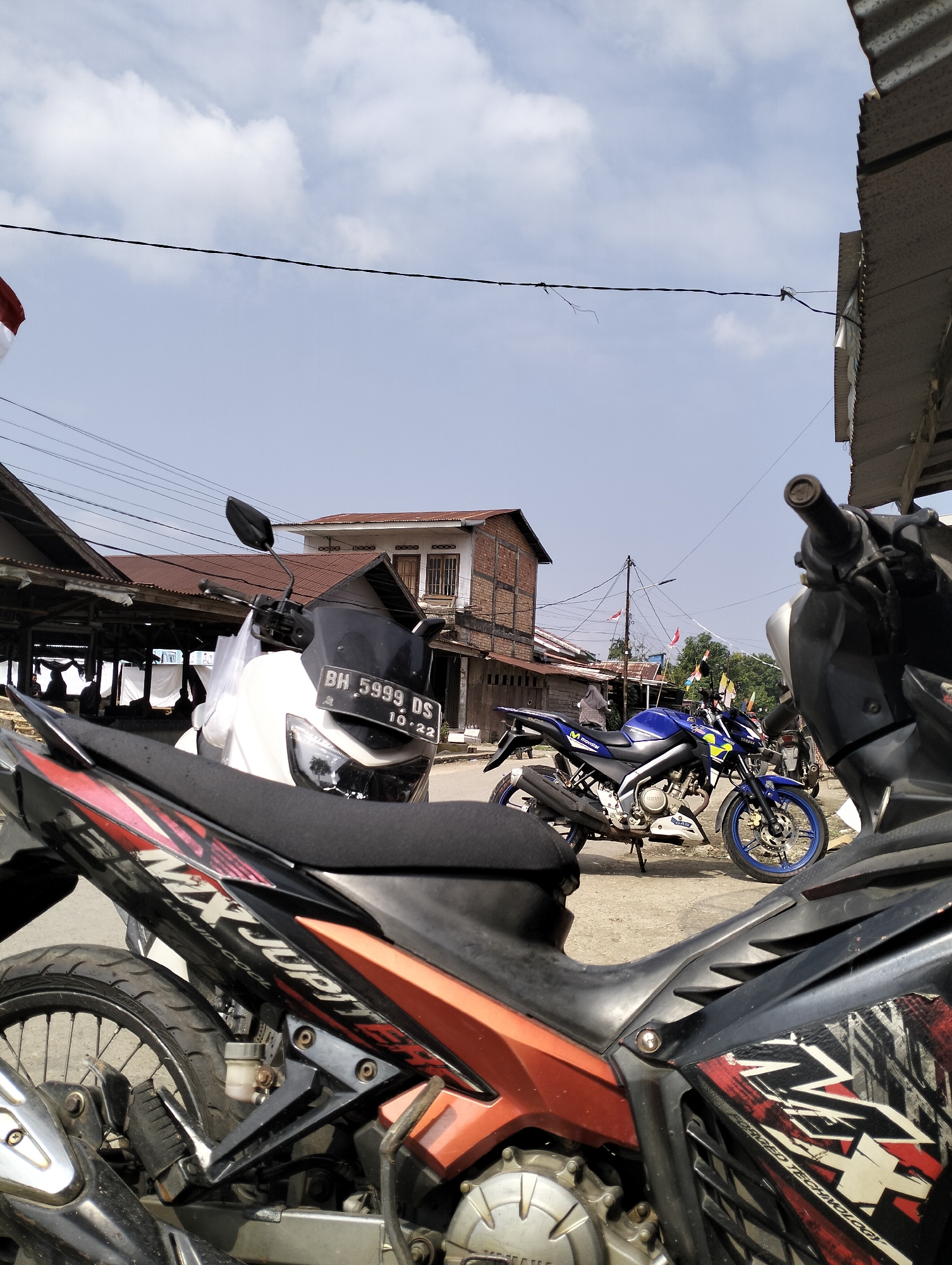 The image depicts a street scene in Serdang Jaya, Indonesia, dominated by parked motorcycles. In the foreground, a black and orange motorcycle is prominently displayed, with its rear wheel and part of its engine visible. Further back, a white motorcycle with a license plate reading "BH 5999 DS" is parked on the asphalt road. To the right of the white motorcycle, a blue Yamaha motorcycle is also visible. The background features several buildings with corrugated metal roofs, some appearing weathered. Electrical wires crisscross the sky, and small red and white flags are strung along a pole in the distance, suggesting a festive or national occasion. The sky is clear and blue, indicating a daytime setting. The overall impression is one of a typical urban streetscape in Indonesia, characterized by the prevalence of motorcycles and the architectural style of the buildings.