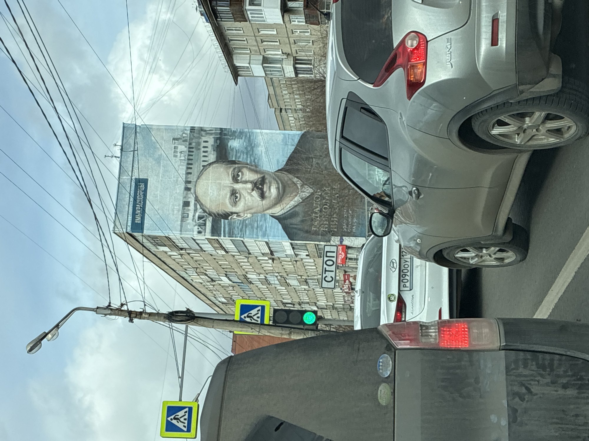 The image depicts a street scene with several vehicles in traffic, seemingly at a stop. A billboard featuring a portrait of a man is visible on the side of a building. The environment appears to be an urban setting with multi-story buildings. A traffic light is present, showing green. A stop sign and a pedestrian crossing sign are also visible. The license plate P090OY 12 on a white car is legible.