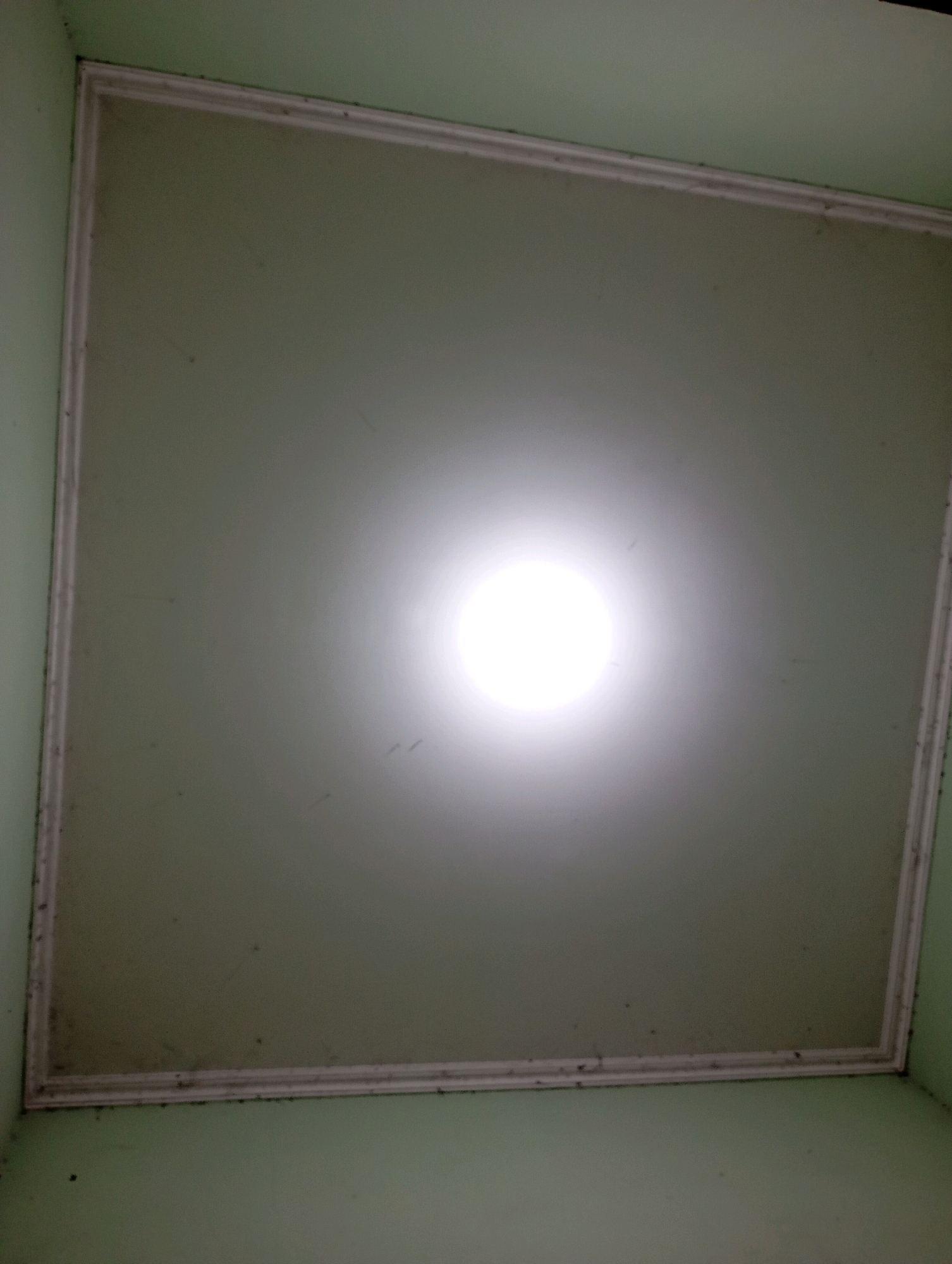 The image shows a close-up, low-angle view of a light fixture in a pale green room. The light fixture is circular and emits a bright white light, creating a halo effect on the ceiling. The ceiling is framed by white molding, and there are some small dark specks visible on the ceiling and molding, which could be dust or dirt. The walls of the room are also pale green. The scene appears to be indoors. There is no indication of the location being in Babelan, Indonesia.