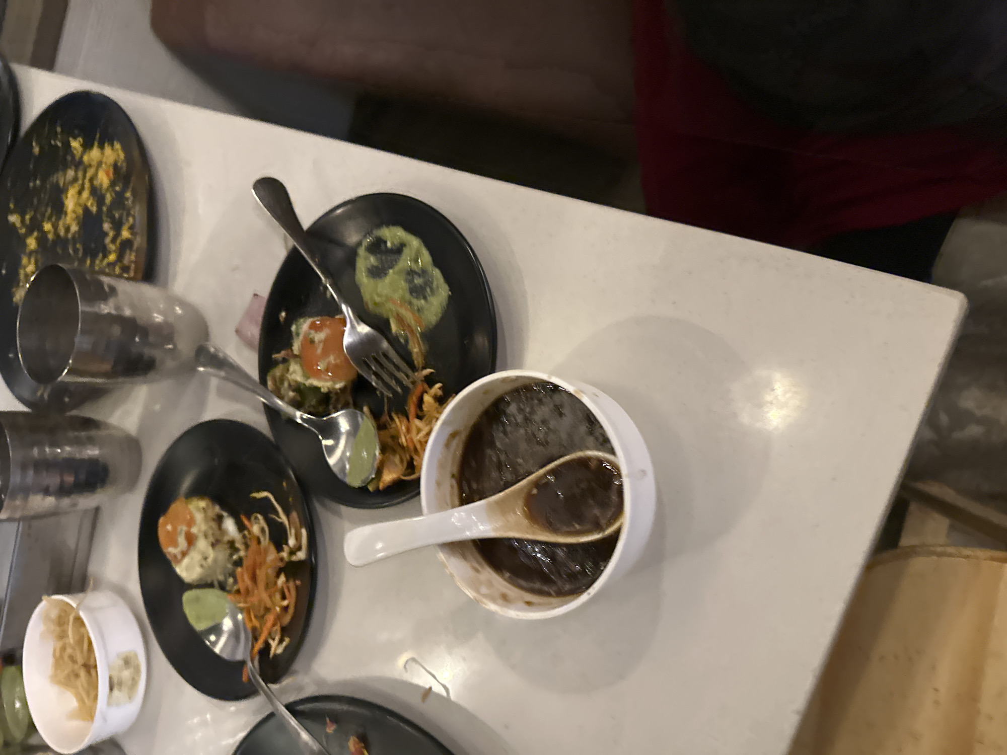 A high-angle shot depicts a table laden with dishes, likely in a restaurant in Kalamb, India. Several black plates hold remnants of food. One plate has food debris mixed with yellow particles. Other plates show half-eaten portions of unidentified appetizers and garnishes. Silverware, including a fork and spoons, sits on or near the plates. A white bowl contains a dark liquid, presumably a sauce or gravy, with a spoon inside. A small white cup holds light-colored food. The table is light-colored, possibly laminate or another type of tabletop material. A person wearing dark clothing and a red garment is partially visible in the background. A beige object is positioned on the lower right edge of the table.