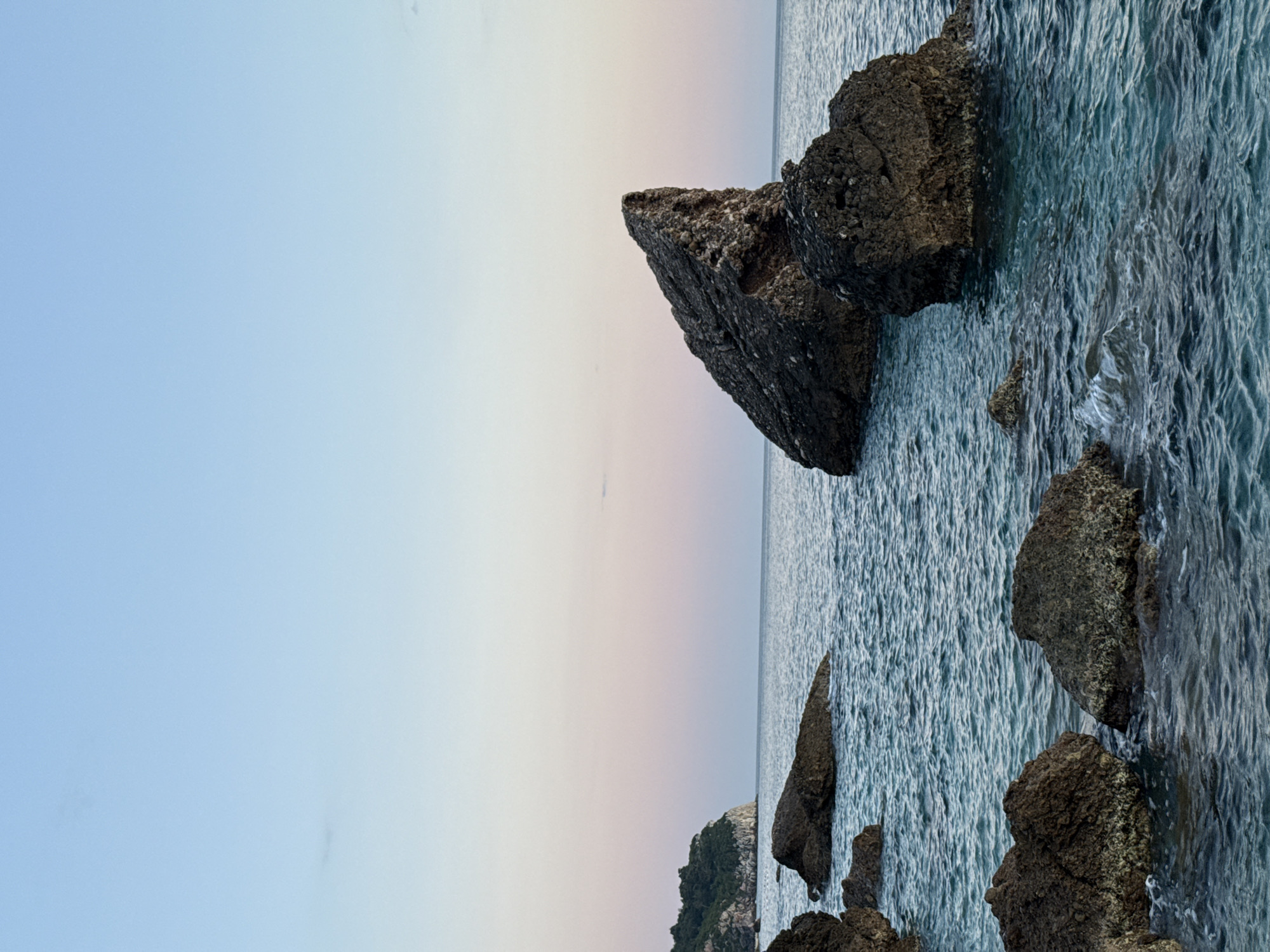 The image shows a view of the sea and rocks. The sky is light blue on the left, fading to a light pink towards the horizon. The sea is a darker blue, with ripples visible on the surface. Several large, dark-colored rocks are present, some protruding from the water. One prominent rock formation appears to be a large, triangular chunk. Smaller rocks are visible near the bottom of the frame. The image has been rotated, with the sky on the left. The location is unknown.