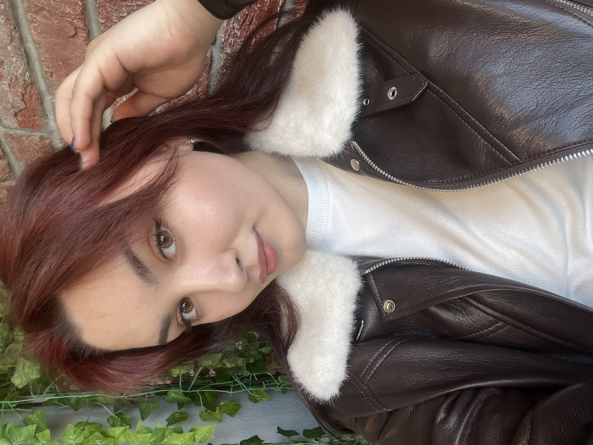 The image shows a woman lying down. She has reddish-brown hair and brown eyes. She is wearing a white top and a brown leather jacket with white faux fur trim around the collar. Her hand is near her hair. The background consists of a brick wall and some green foliage. The location is unknown and cannot be determined from the image.