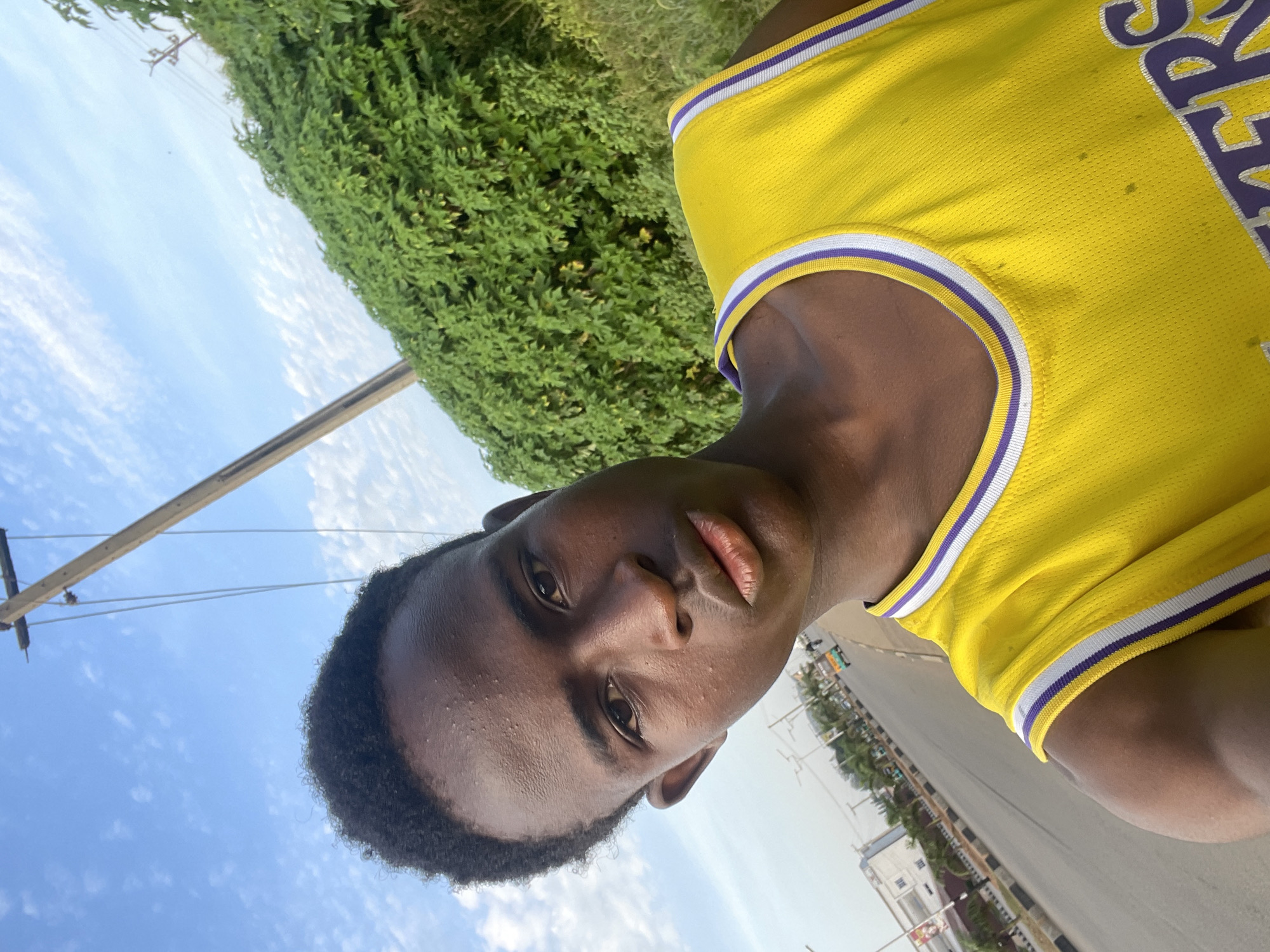 A young Black male, facing the camera, stands outside in what appears to be a city setting. He is wearing a bright yellow basketball jersey with purple and white trim. His hair is short and dark. Behind him, a wooden utility pole with wires stretches upwards against a bright blue sky dotted with wispy white clouds. To the right, a dense, green hedge forms a visual barrier. In the background, a road curves to the left, lined with buildings and palm trees. A few vehicles, including what looks like a tricycle taxi, are visible on the road. The overall impression is an outdoor, daytime shot in an urban environment. The context provided suggests this location is the city of Jos, Nigeria.