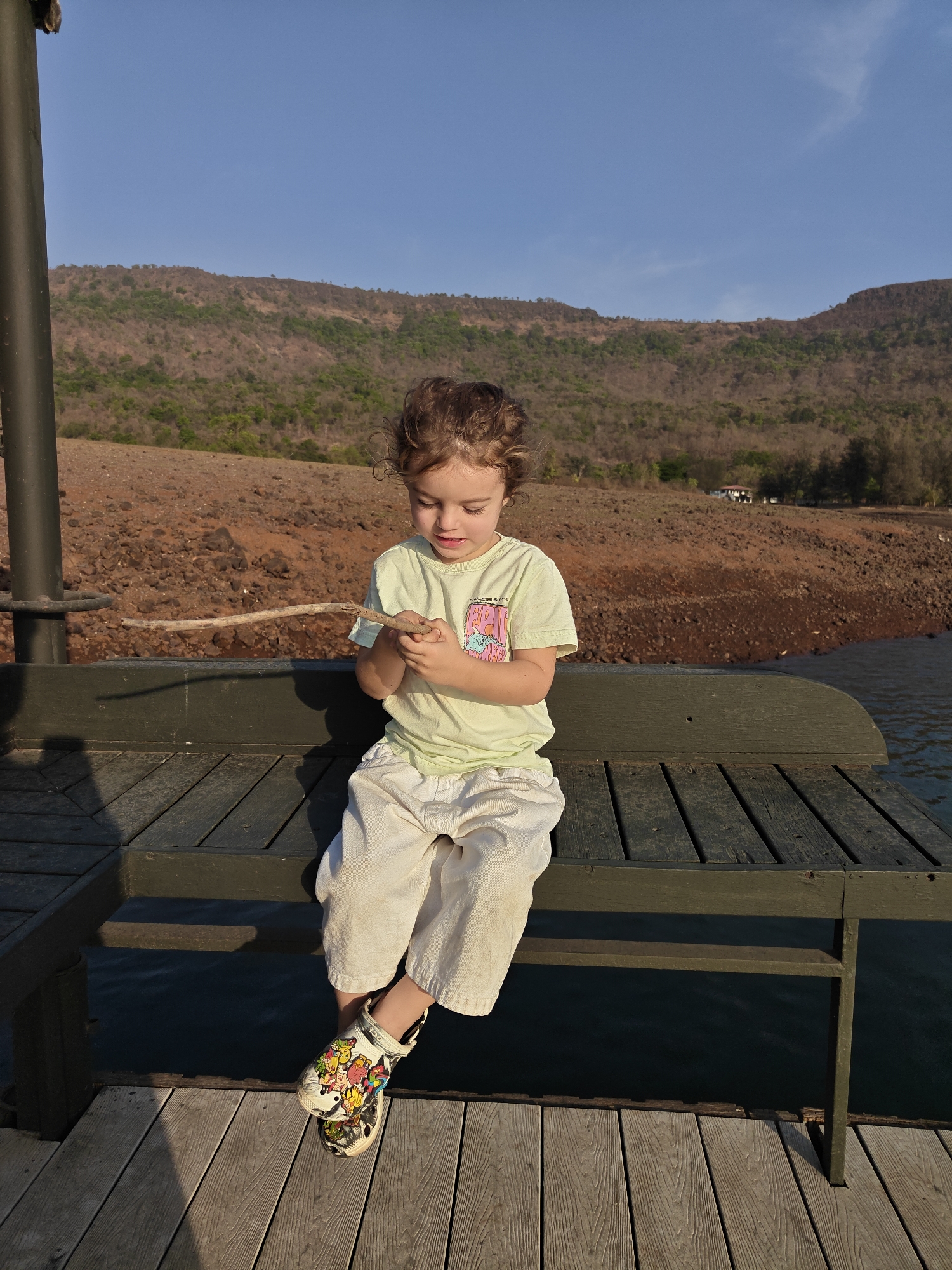A young child with curly brown hair is sitting on a wooden bench at what appears to be a dock or pier. The child is wearing a light green t-shirt, white pants, and colorful Crocs shoes. The child is holding a small twig or stick in their hands and is looking down at it with focused attention. Behind the bench, there is a body of water. Beyond the water is a landscape of rocky terrain and mountains. The sky is blue with some sparse clouds. A dark metal pole is visible to the left of the child. The scene is bathed in warm sunlight, suggesting it is either morning or late afternoon. This image was taken in Kalote Mokashi, India.