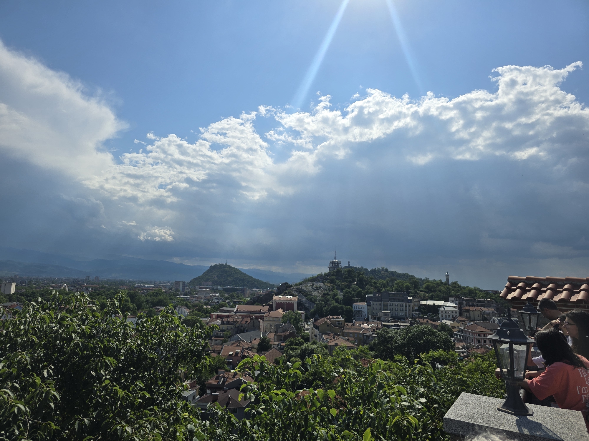 Here is a description of the image:
The image is a high-angle, outdoor daytime shot of a city landscape, dominated by a partly cloudy sky with visible sun rays. The city is likely Unknown, Unknown, based on the architectural style. The scene features buildings with reddish-brown tiled roofs mixed with more modern structures, all surrounded by lush green trees. In the midground, two hills are visible. One is covered with trees and has a telecommunications tower on top, and another is slightly smaller. In the foreground, there is foliage, a concrete ledge, and several individuals with dark hair are situated near antique-style street lamps.