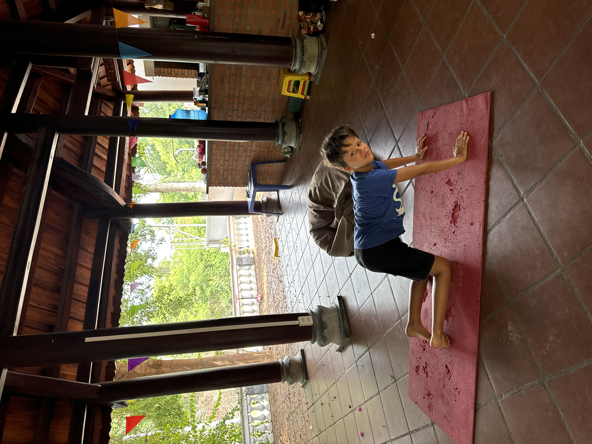 A young boy is on his hands and knees on a red yoga mat, on a tile floor, in what appears to be an open-air covered pavilion in Mũi Né, Vietnam. The boy is wearing a blue t-shirt and black shorts. The pavilion has a wooden roof and large wooden pillars. In the background, there are trees and foliage, and a brick wall. There are also colorful flags strung across the pavilion.