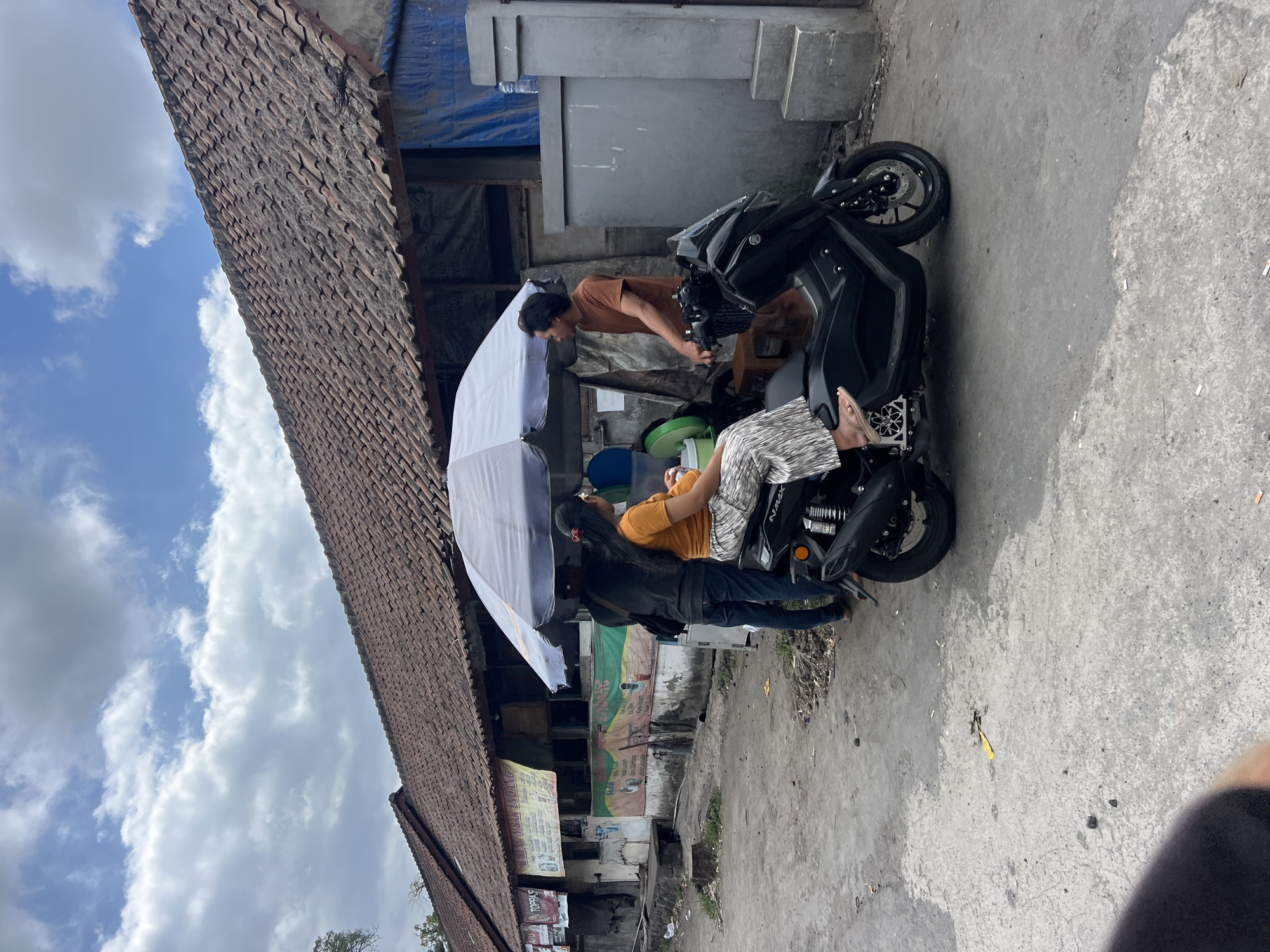 In Ubud District, Indonesia, an outdoor scene unfolds under a cloudy sky, featuring a street-level view of a building with a tiled roof and a gray wall with steps. A black scooter is parked on a dusty road, with a woman in a tan shirt and patterned pants seated on it. Beside her, a man in a tan shirt stands, appearing to interact with her or the goods under a white umbrella that shades a small stall. Another person, wearing a black shirt and blue jeans, stands behind the woman on the scooter. The stall appears to be selling various items, with banners and signage visible on the building behind. The ground is a mixture of dirt and what appears to be cracked concrete, with some scattered debris.