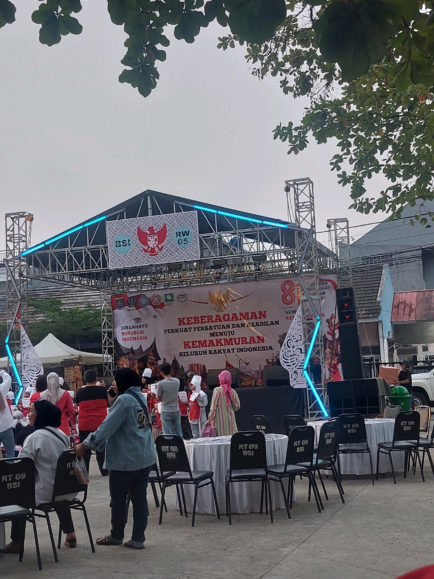 A daytime, eye-level outdoor shot captures a community gathering in front of a stage in Depok, Indonesia. The sky is overcast. A stage structure with a dark roof and blue LED lighting is set up, displaying banners with Indonesian text and logos. Among the logos, a prominent Garuda Pancasila emblem is visible, alongside "BSI" and "RW 05". Below the stage, round tables covered with white tablecloths are arranged, each with several black chairs bearing the "RT 09 BSI" insignia. A crowd of people, including men, women, and children, are present. Some are seated at the tables, while others stand, observing the stage or interacting amongst themselves. Many individuals are wearing traditional Indonesian attire, including headscarves and peci caps. The ground is paved with concrete. Trees with green foliage overhang the scene from the top. In the background, residential buildings and a white car are visible.