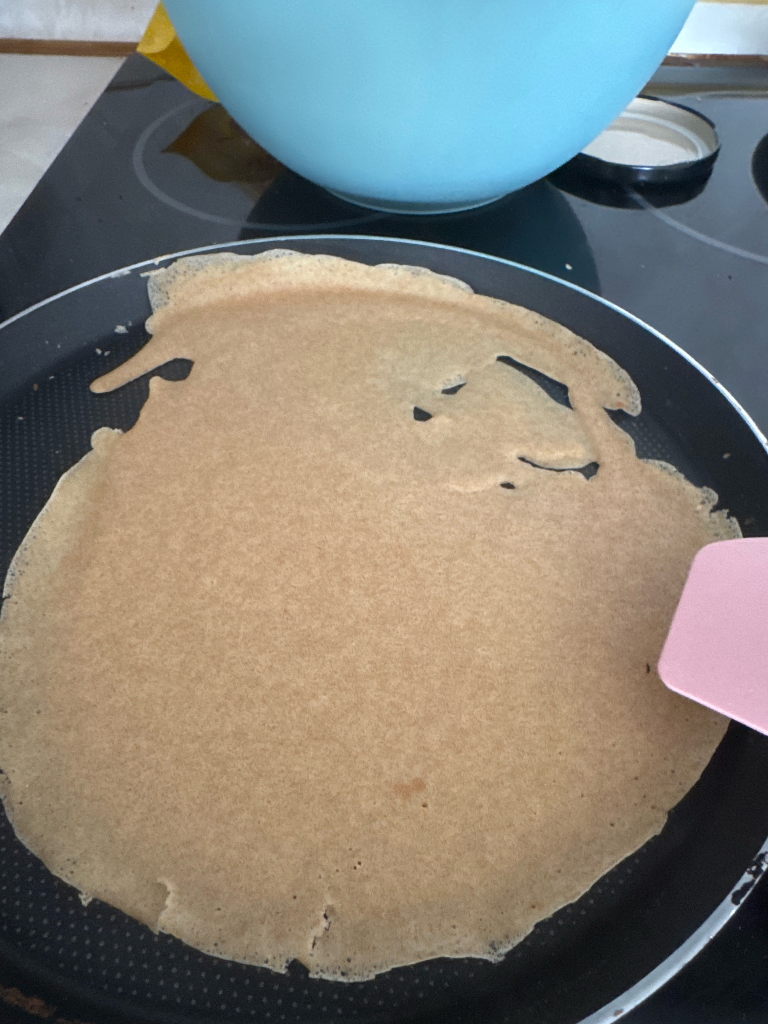 An overhead shot shows a light brown crepe cooking in a black non-stick pan on a stovetop. The crepe is irregularly shaped, with a few small holes. A pink spatula is visible on the right side of the pan, partially covering the edge of the crepe. In the background, a blue bowl sits on the stovetop next to a black lid. The scene appears to be in a kitchen. The city of Sofia, Bulgaria, is not visible or referenced in this image.