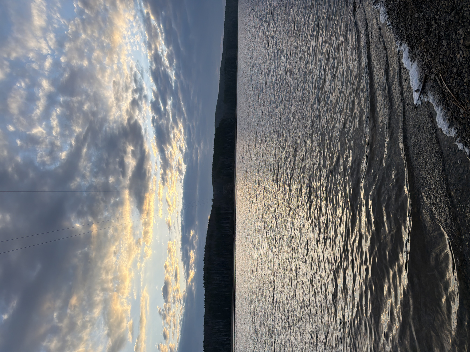 The image depicts a landscape with a body of water, likely a lake, and the sky. The left side shows a cloudy sky with hints of sunlight breaking through. The clouds vary in color from white to gray, with some illuminated by a warm, golden light. Power lines are visible against the sky. In the center of the image, a dark, forested shoreline separates the sky from the water. The right side displays the surface of the water, reflecting light and showing small ripples or waves. The shoreline is visible at the bottom right, with small rocks and possibly some foam or debris along the edge. The location is possibly Chaykovsky, Russia based on the geographical features and landscape elements, though the image does not definitively confirm the location.
