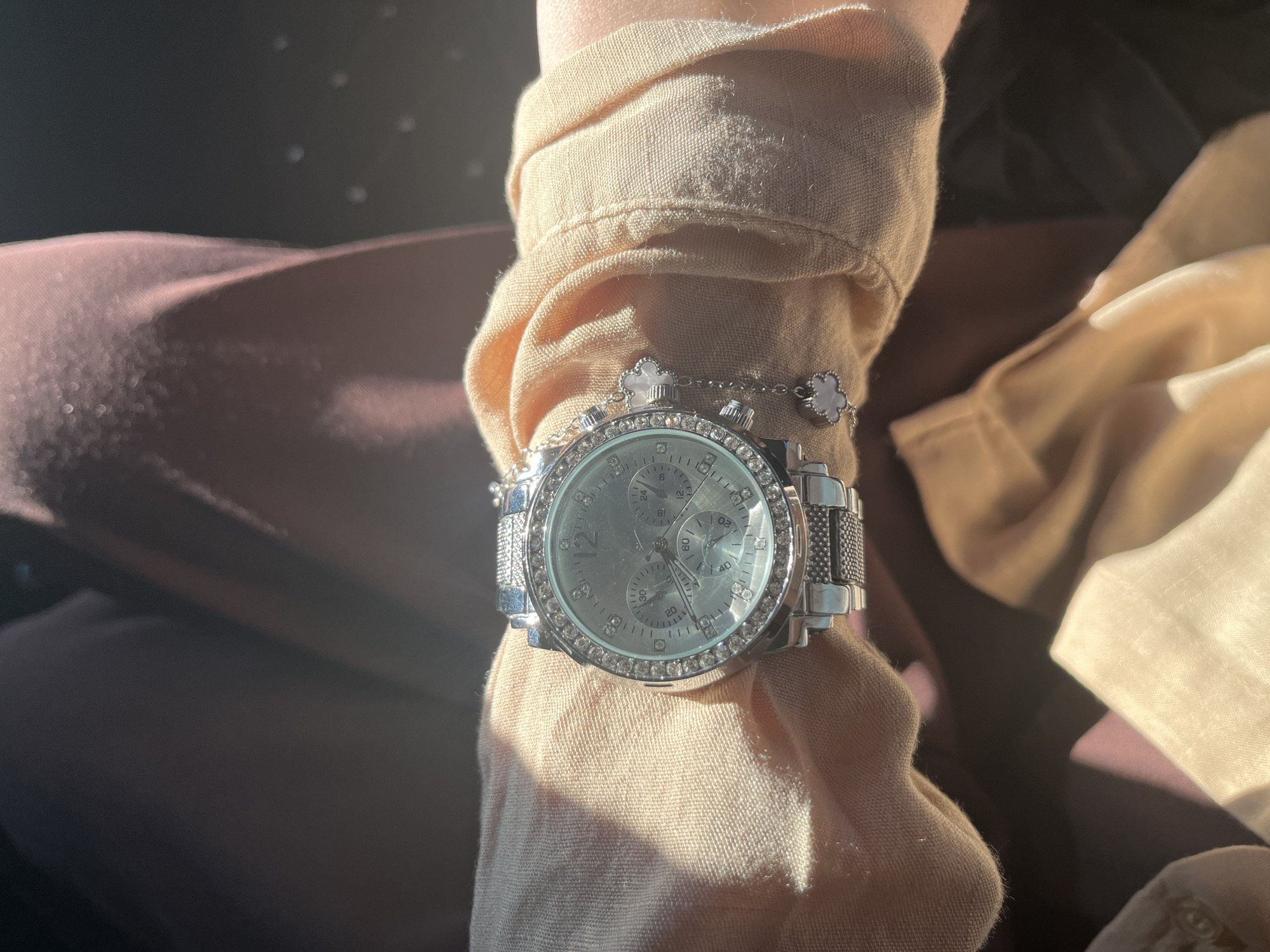 An individual is wearing a silver, jeweled wristwatch on their left wrist. The watch has multiple subdials and a jeweled bezel. A bracelet with small, clover-shaped charms is attached to the watch. The person is wearing a light-colored, possibly linen, shirt with puffed sleeves. The background is out of focus, with hints of dark clothing or fabric in shades of maroon and brown. The lighting suggests an indoor setting with sunlight filtering in from the side, creating bright highlights and shadows. There is no discernible information about the location being in Загородный, Russia.