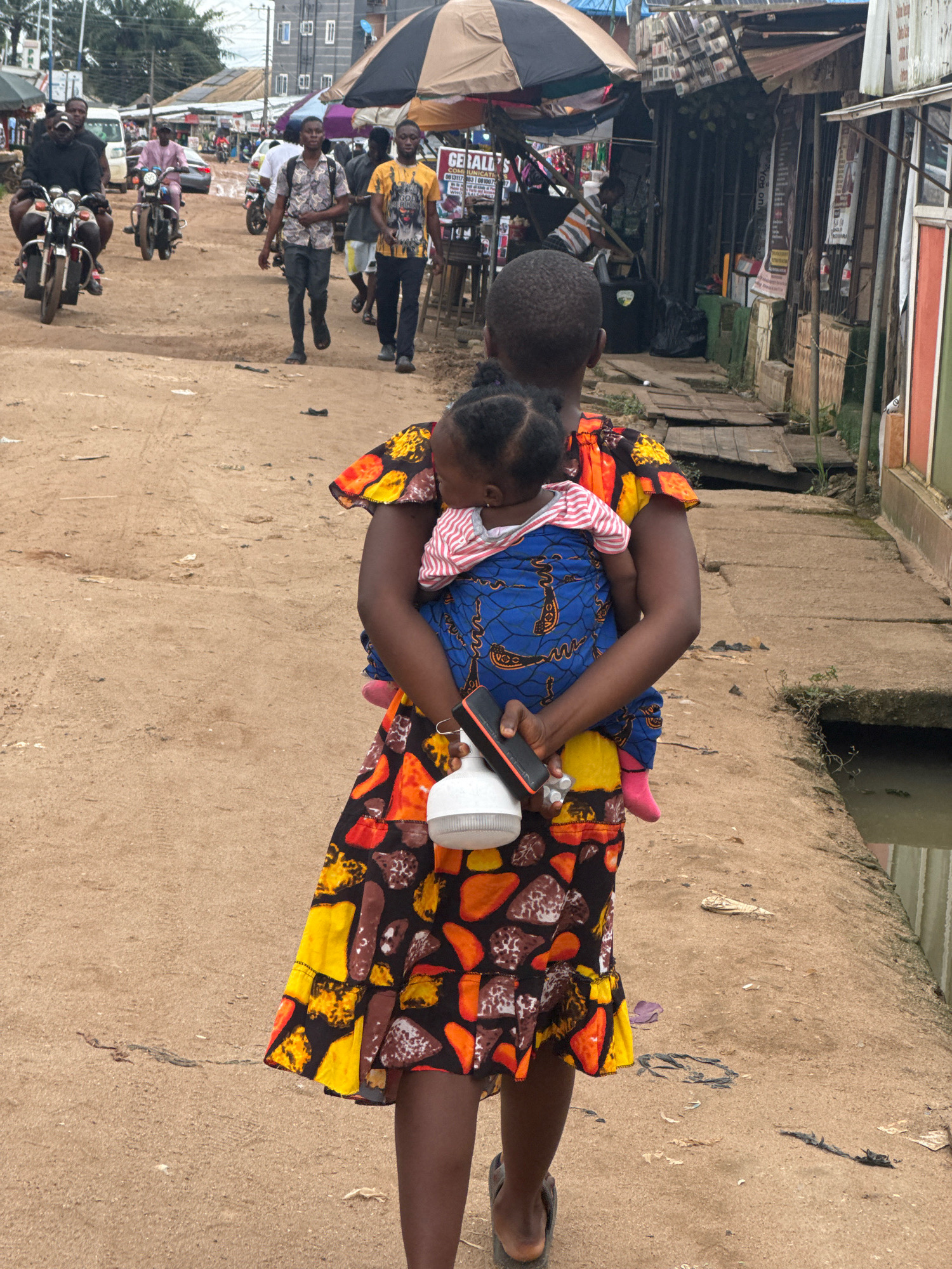In Ihiagwa, Nigeria, a woman carries a baby on her back while walking on a dirt road. She is wearing a colorful, patterned dress and carrying a white object with an orange handle in her right hand. The baby is wrapped in a blue cloth with a yellow and white pattern. In the background, several men are riding motorcycles and walking. Shops and stalls line the right side of the road. The ground is dusty and uneven, with scattered debris. A narrow channel of water runs alongside the right edge of the road. The overall scene depicts a bustling market area in a Nigerian town.