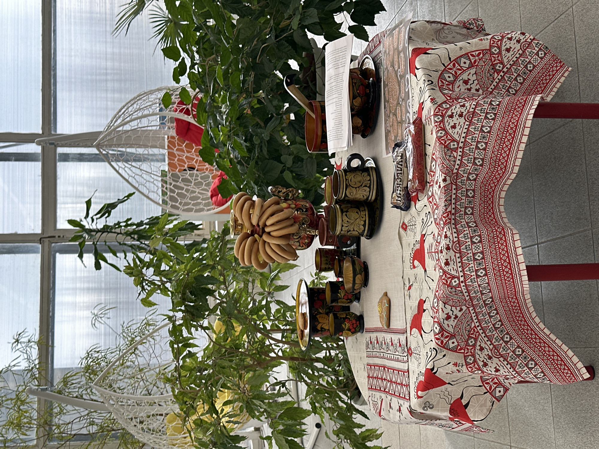 Captured indoors in what could be a botanical garden or atrium, the scene presents a table adorned with a patterned tablecloth featuring shades of red and white. The table serves as a display for a traditional tea set, including mugs, a teapot, and a stack of bagels or donut-shaped pastries arranged around the teapot's lid. Lush greenery, including small trees and plants, surrounds the table, creating a vibrant backdrop. Through the glass panels of the structure, hints of the outside world can be seen. A white, hanging chair swing is visible in the background.