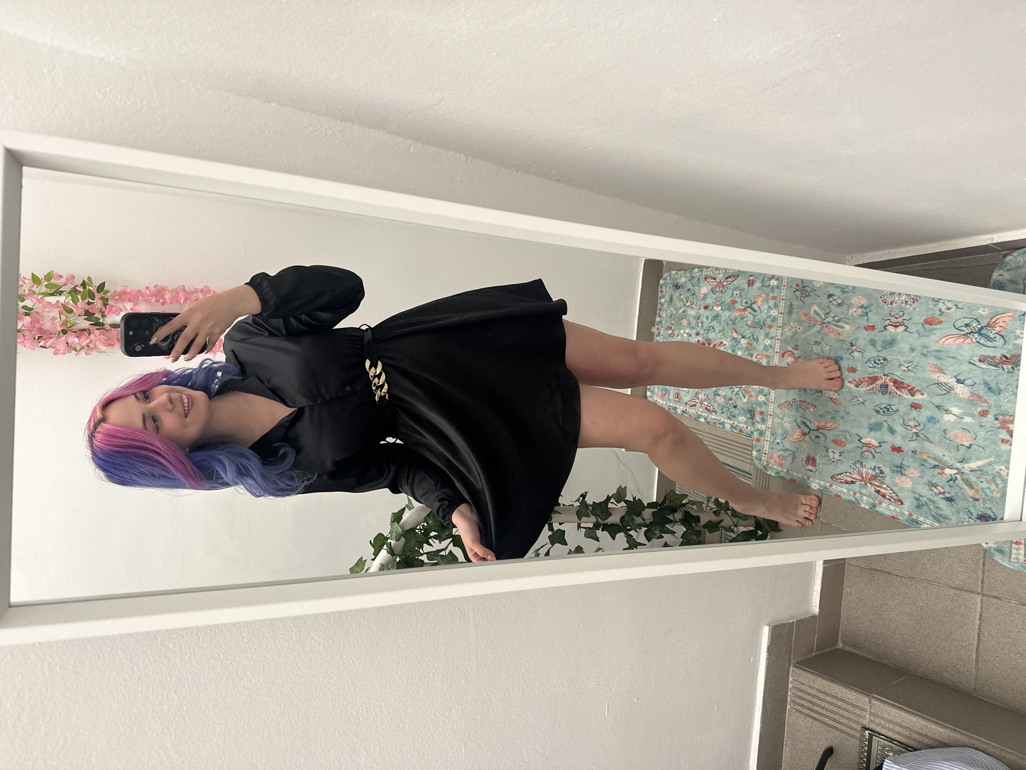 This image captures a mirror selfie of a young woman in an indoor setting, likely in the City of Zagreb, Croatia. The photograph is oriented vertically, with the woman standing upright, but the image itself is rotated 90 degrees counter-clockwise.
The primary subject is a young woman, seen from head to toe, taking the photo with a black smartphone held in her right hand. She has striking, long hair colored in vibrant shades of pink and purple/blue. She is smiling broadly, conveying a cheerful and confident mood.
She is dressed in a long-sleeved black mini-dress, which is cinched at the waist by a distinctive gold chain belt. Her bare feet are visible, and she appears to be standing casually for the selfie.
The setting visible in the mirror's reflection includes a portion of a light blue rug adorned with colorful patterns of butterflies, beetles, and floral motifs. Beside the mirror, a string of pink artificial flowers is draped on the wall, and green artificial ivy is visible near the bottom of the frame, hinting at decorative elements within the room. The walls reflected in the mirror are plain white. Outside the mirror's frame, a tiled floor, possibly grey or light brown, is visible, suggesting the mirror might be in a hallway, bedroom, or dressing area adjacent to a bathroom.
There is no visible text in the image. The time of day is not discernible due to indoor lighting. The activity is purely a personal photo-taking moment.