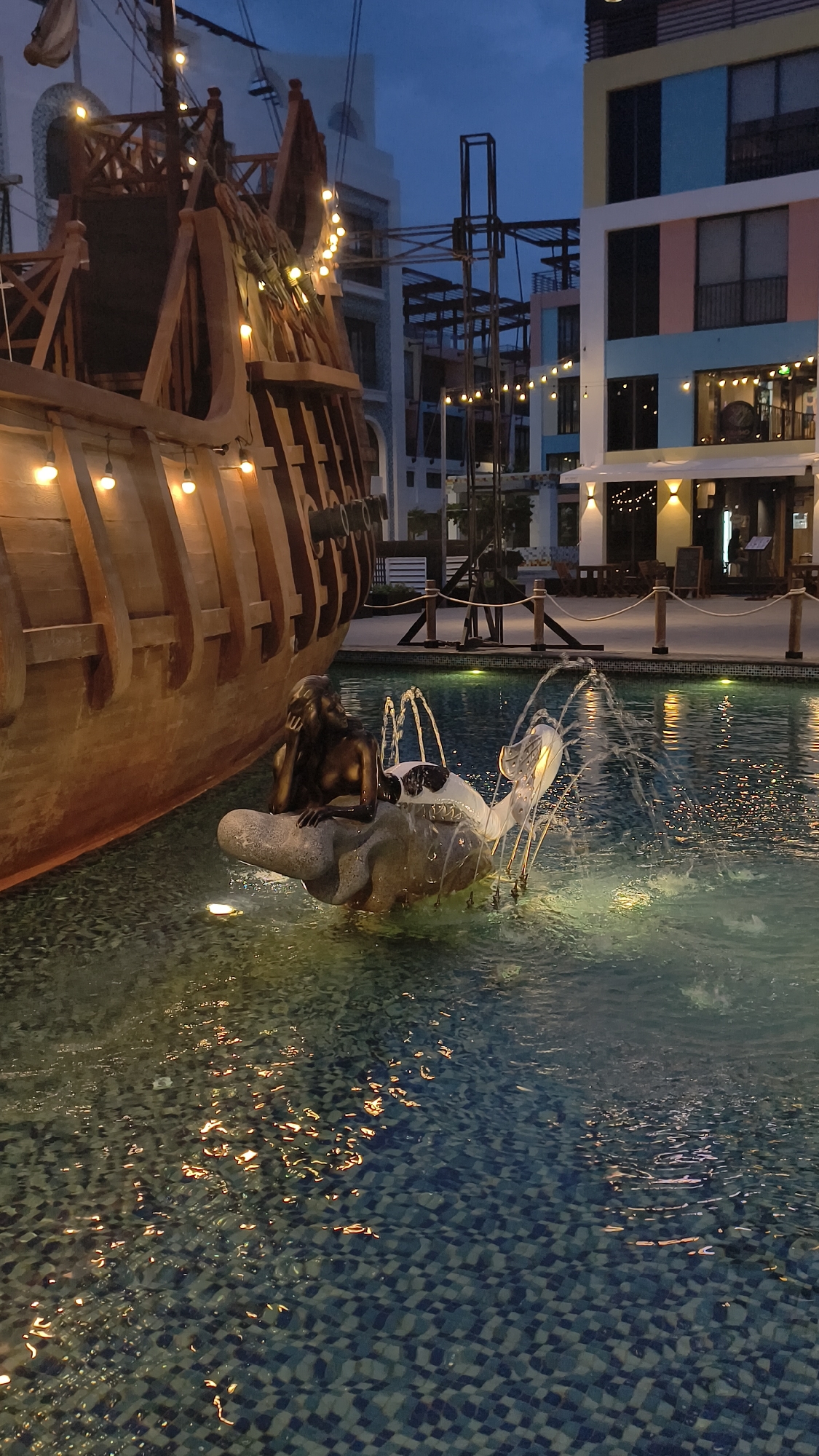 The image depicts an outdoor scene at dusk in Nha Trang, Vietnam. Dominating the left side is a large, wooden replica of a ship's hull, adorned with a string of warm-toned lights. In the foreground, a pool of water features a bronze statue of a mermaid resting on a shell-like structure, with water jets creating a fountain effect. Behind the ship, a multi-story building with a modern architectural style and colorful facade is visible, also decorated with lights. The overall ambiance suggests a recreational or entertainment area, possibly a waterfront promenade or a themed attraction.