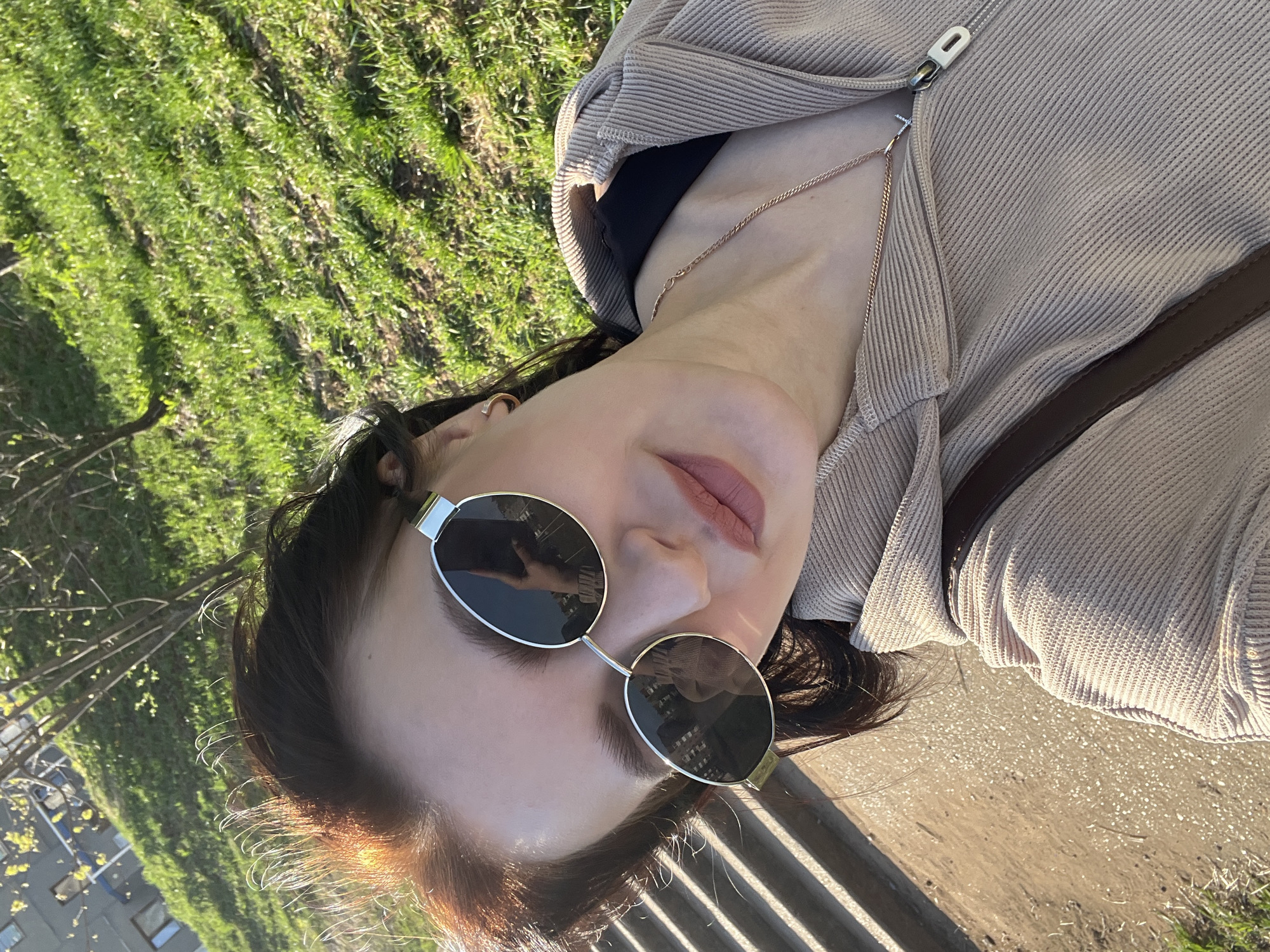 A woman is pictured outdoors in Markovskii, Russia, wearing sunglasses and a beige zip-up hoodie with a brown bag strap visible. She has a gold necklace. The background shows green grass, trees, a building with windows, and a bench with shadows cast across it. The woman is possibly sitting or reclining on the bench.