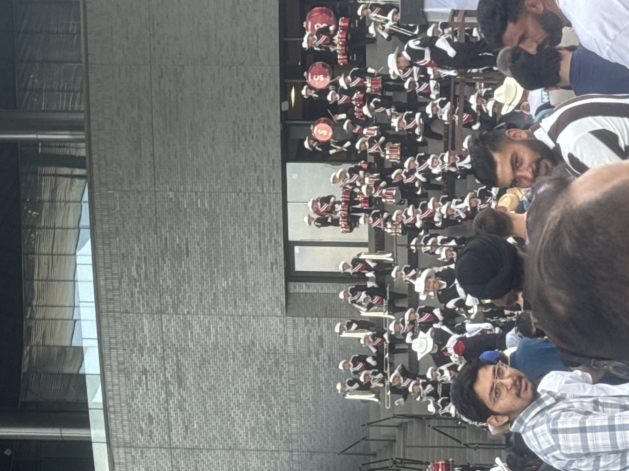 This is a vertical, slightly low-angle, full shot photograph of a marching band performing outdoors in front of a brick building. The band members are dressed in black and white uniforms with white hats, and many are holding instruments like drums and tubas. They are arranged on tiered steps. In the foreground, several members of the audience are visible, some looking towards the band and others looking in different directions. One man in the foreground is wearing glasses and a plaid shirt. The brick building behind the band appears to be modern, with large glass windows on an upper level. The overall atmosphere suggests a public event or performance. This scene is located at the Calgary Stampede grounds in Calgary, Canada.