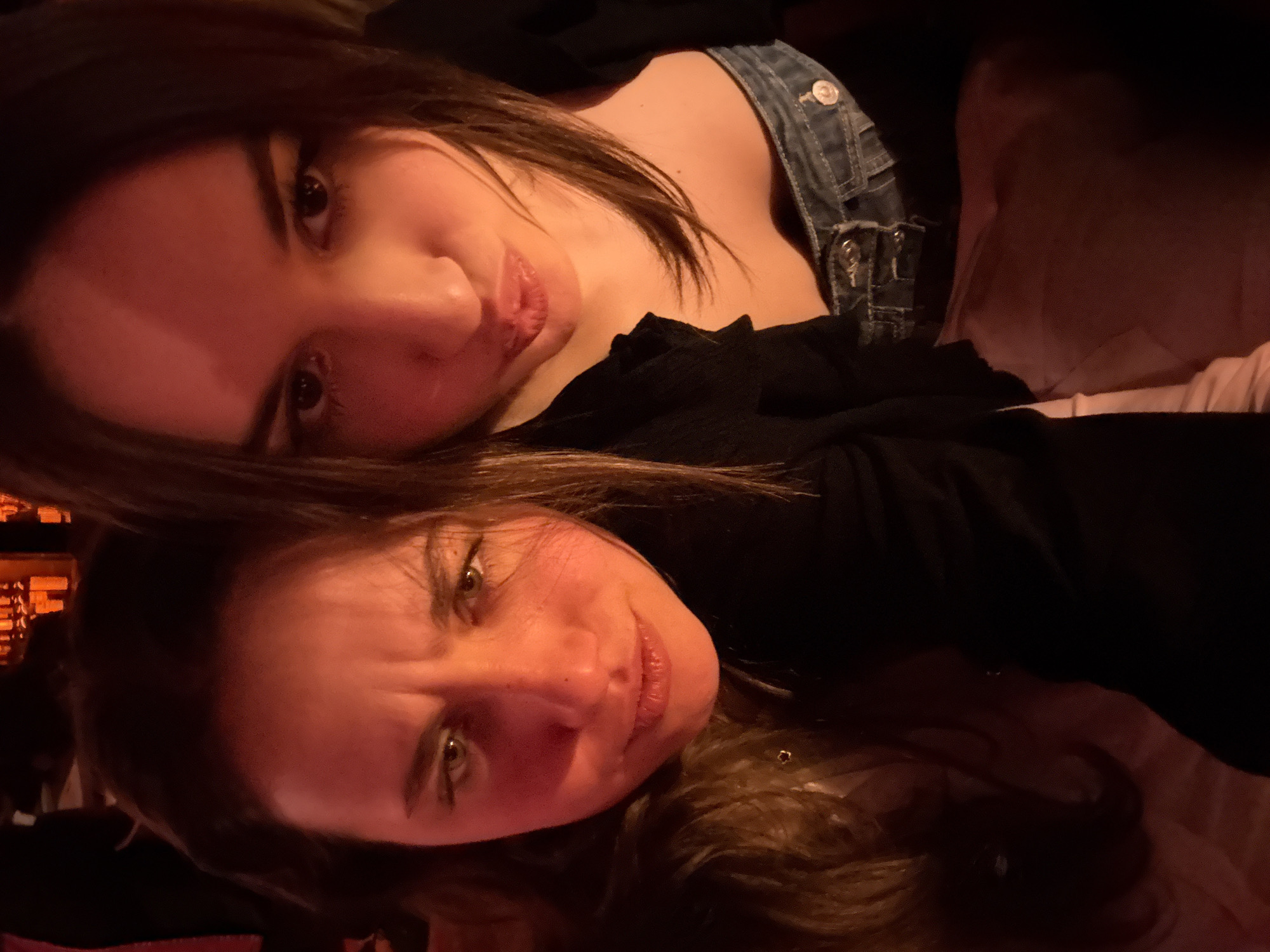 The image displays two young adult females in a close-up portrait, captured in a dimly lit indoor environment with a pervasive reddish-orange light source. The background is out of focus, revealing faint amber-lit shapes consistent with bottles or decorative objects on shelves, suggesting a bar or restaurant setting.
The individual positioned on the left, with long dark hair, presents a playful scowl, characterized by a furrowed brow, narrowed green eyes, and a slightly upturned mouth. She wears a dark-colored top.
The individual on the right, also possessing long dark hair, adopts a pouting expression with lips pursed forward. She is attired in a dark outer garment over a lighter-colored inner top, with blue denim jeans visible at her waist.
While the prompt indicates the location is Milan, Italy, specific visual identifiers within the image itself do not confirm this city or any precise location within it.