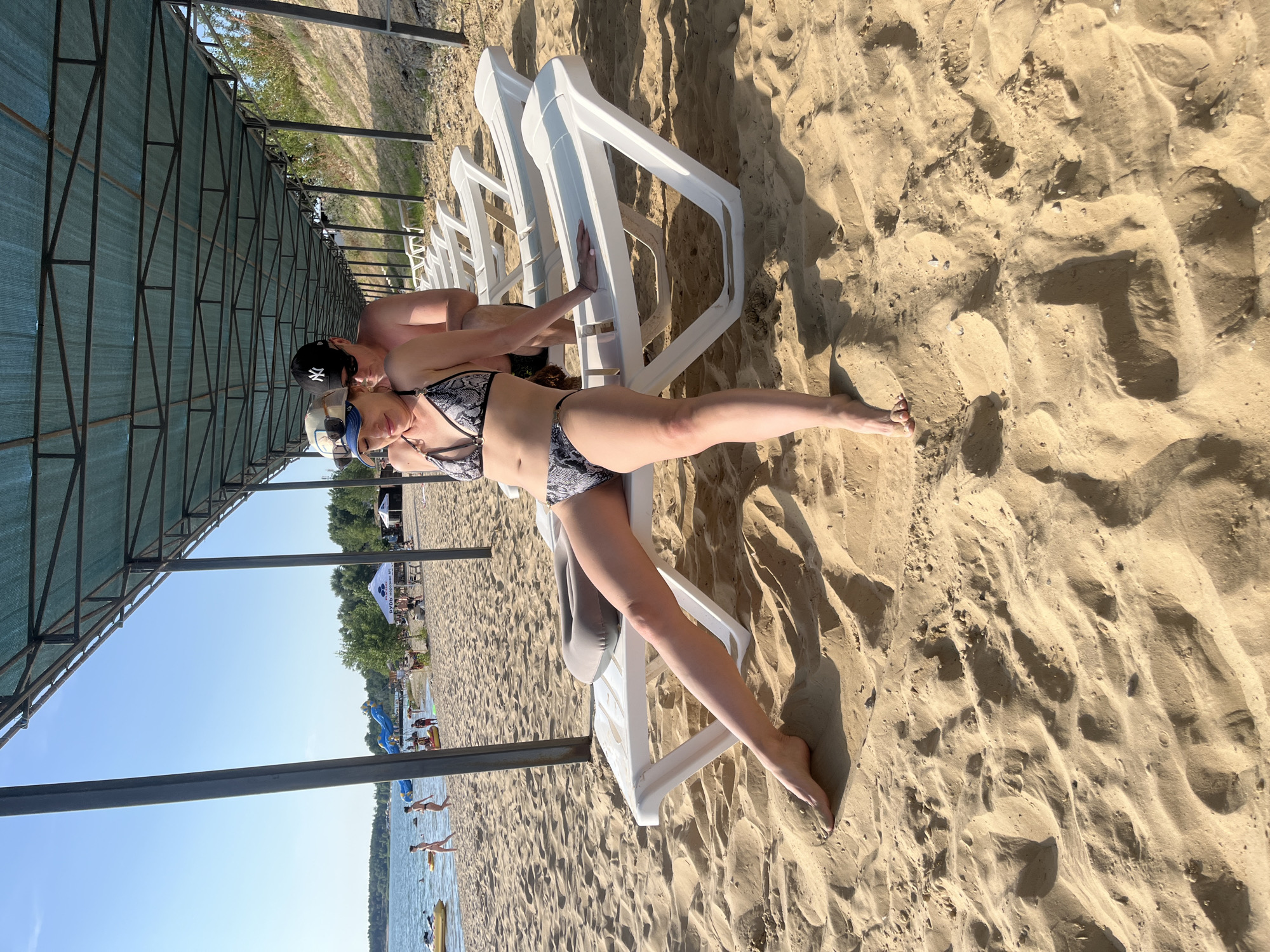 The image shows two people on a sandy beach. One person is lying on a white lounge chair, wearing a black and white patterned bikini and sunglasses. The other person is partially visible behind the lounge chair, wearing a black baseball cap. They are under a shaded structure with a green canopy and metal supports. The beach is covered in sand with ripples and shadows. In the background, there is a body of water with people swimming and a few yellow inflatable devices. Trees and a clear blue sky are visible beyond the water. The scene appears to be a sunny day at a beach resort or public beach. There is no visible text in the image.
