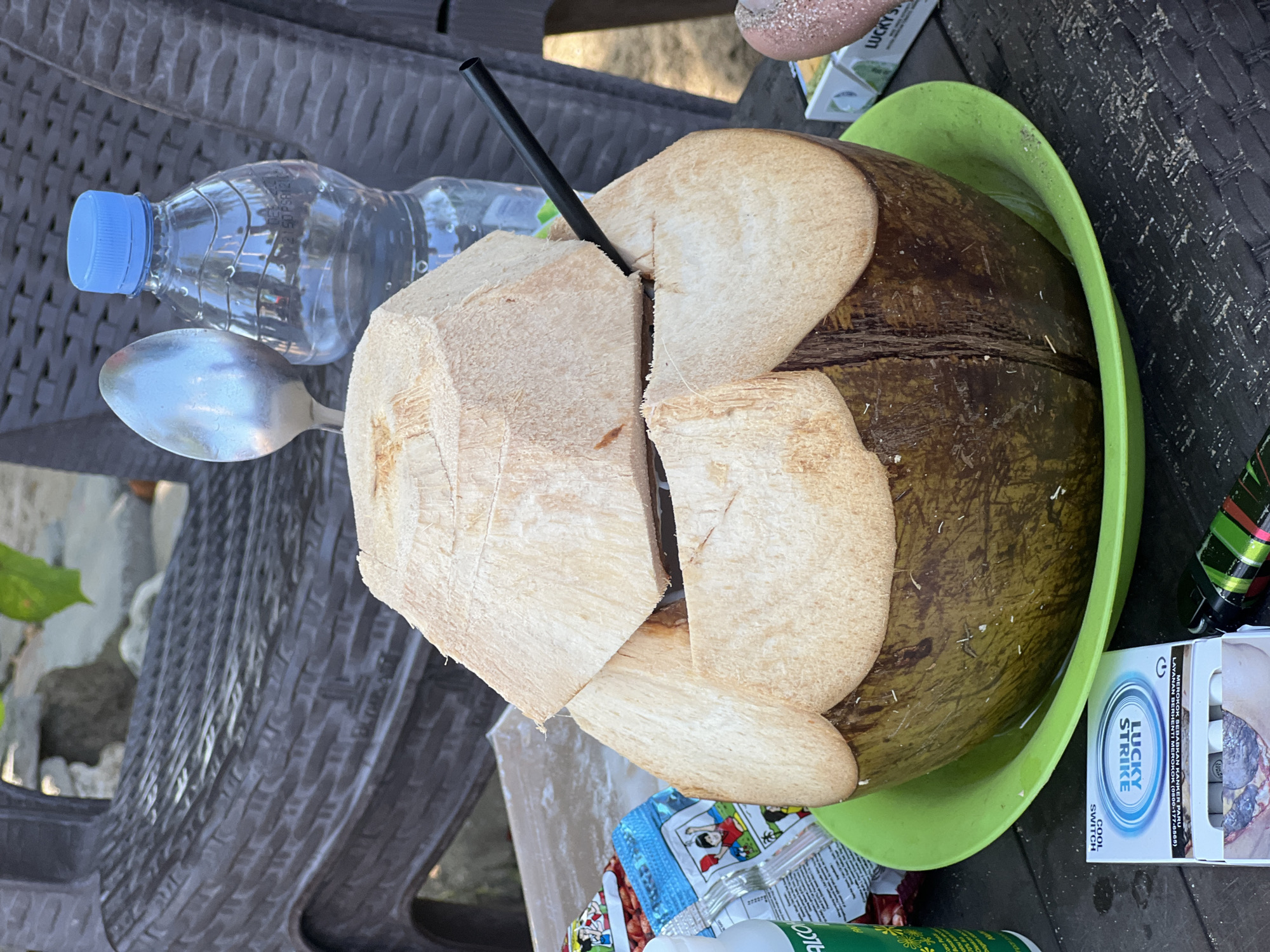 This image depicts a close-up view of a large, opened coconut placed in a bright green plastic bowl on a dark, textured surface, likely a table. The coconut has been cut from the top, with the lid partially removed and resting on its side, revealing the white flesh and liquid inside. A black straw is inserted into the coconut. To the left of the coconut, a partially visible package with colorful graphics and text is present, along with a white bottle and a silver spoon. In the background, a dark-colored woven chair is visible, along with a clear plastic water bottle with a blue cap. To the right of the coconut, a cigarette pack labeled "LUCKY STRIKE" is visible, along with a lighter. The scene appears to be set outdoors, possibly at a beachside establishment in Kuta, Indonesia, given the casual setting and the presence of a coconut drink.