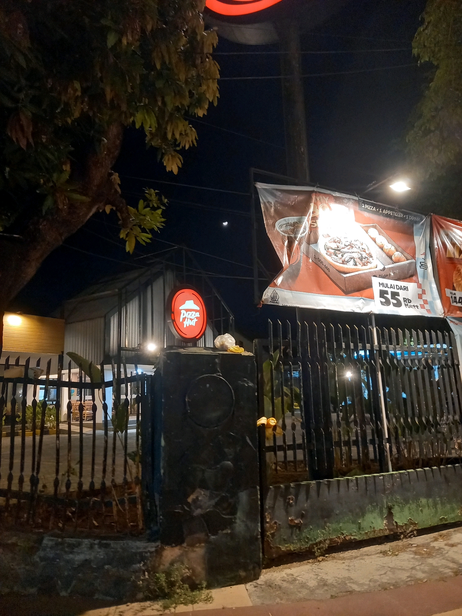 A nighttime, eye-level shot captures the exterior of a Pizza Hut restaurant in Pamekasan, Indonesia. The building features a corrugated metal roof and a yellow-toned exterior wall. In front of the building, a black metal fence with decorative elements encloses the property.
Dominating the scene is a large, illuminated red circular sign with the "Pizza Hut" logo. To the right of the sign, a large banner advertises a pizza meal deal, displaying images of pizza and appetizers, along with pricing information in Indonesian Rupiah. The banner reads "MULAI DARI 55.000 RB Nett," indicating a starting price of 55,000 Rupiah.
A large tree with dark foliage is visible on the left side of the image. The sky is dark, with a faint crescent moon visible. Streetlights and interior lights from the restaurant illuminate the scene. The foreground shows a concrete pathway and a low wall. There are no people visible in the image.