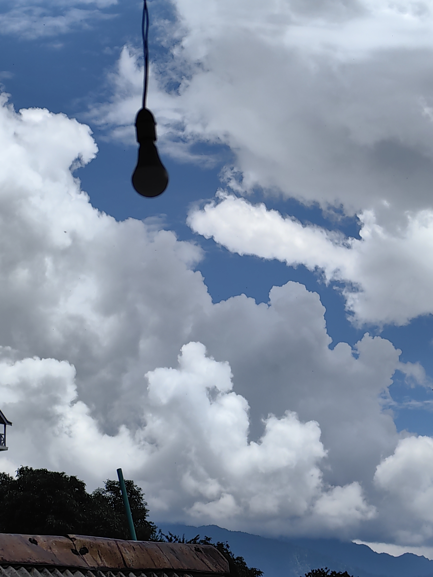 A low-angle shot captures a silhouette of a lightbulb hanging from a dark string against a bright blue sky filled with fluffy white and gray clouds. The bottom of the image shows a section of a rusty corrugated metal roof, some dark green trees, and a distant mountain range. The scene is situated in Vashisht, India.