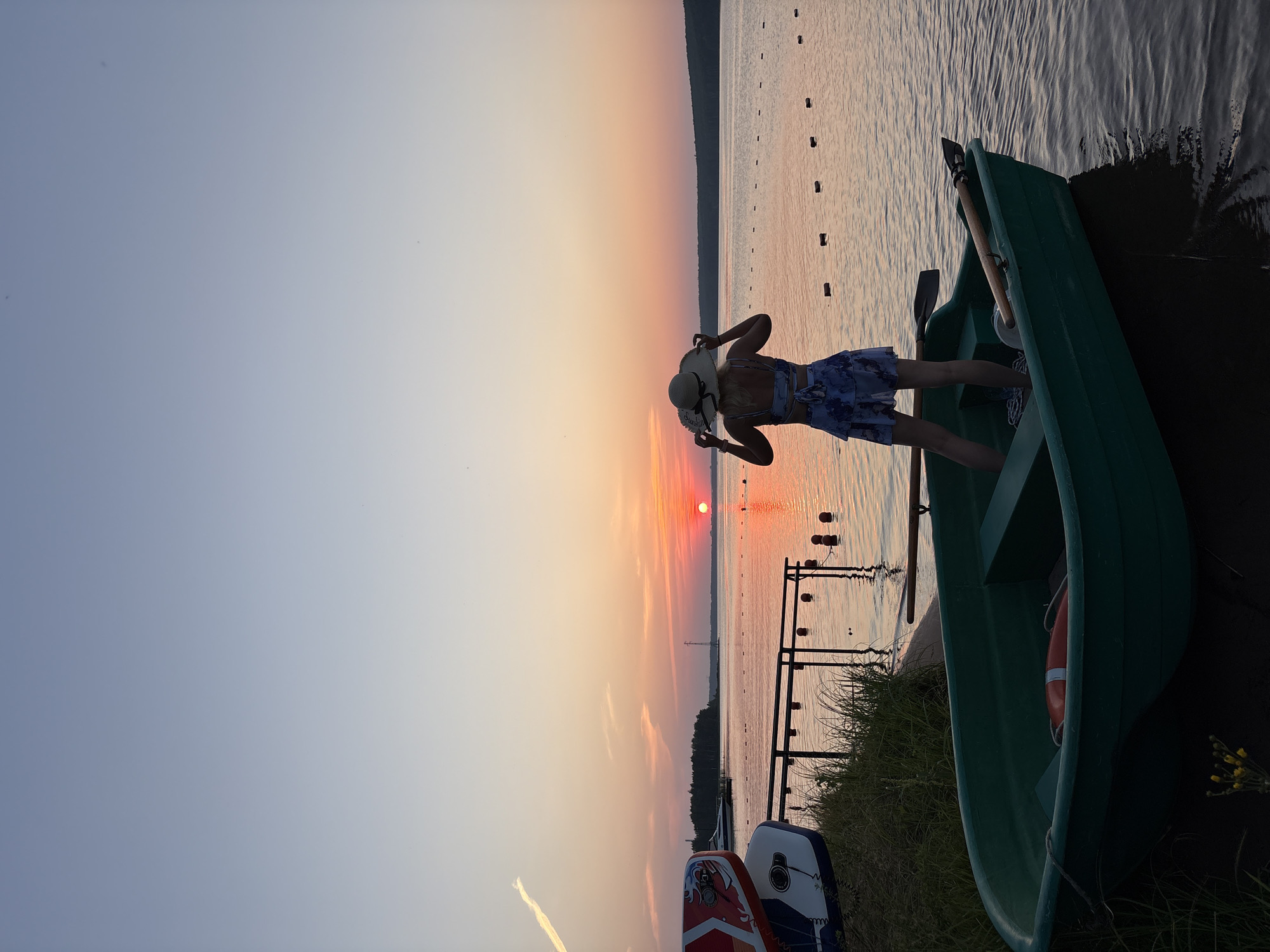 A woman wearing a straw hat and a blue floral print dress stands in a green rowboat at sunset. She is facing away from the camera, looking towards the horizon where the sun is setting over a body of water. The sky is a gradient of pale blue and orange. In the foreground, the rowboat is partially submerged at the edge of the water, with grass and other boats on the shore behind it. There are buoys floating in the water, marking a path or boundary. The location is the Kama River in Chaykovsky, Russia, known for its scenic sunsets.