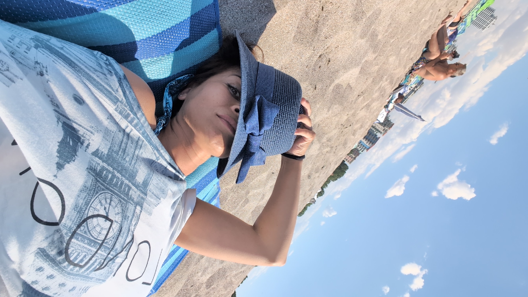 In Constanța, Romania, a woman wearing a blue hat with a bow and a t-shirt with a city skyline print reclines on a blue and white striped mat on a sandy beach. She is lying on her back, holding the brim of her hat with her right hand. In the background, other beachgoers are visible, along with buildings and trees, under a partly cloudy blue sky.