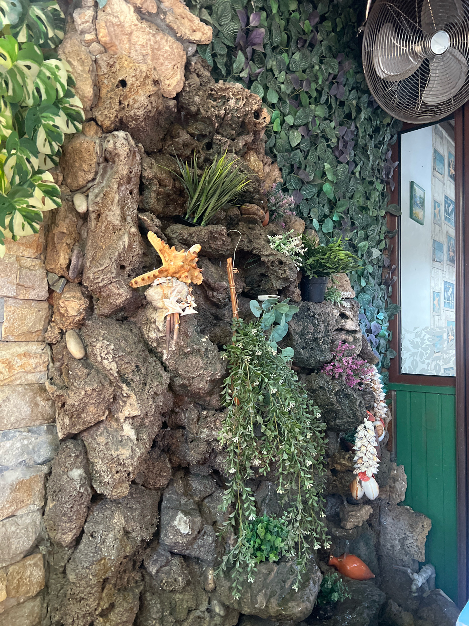 The image depicts a decorative rock wall with an assortment of artificial plants and seashells. A prominent starfish decoration is positioned on the left side of the rock formation. To the right, a metallic fan is visible, and behind it, a wall covered with green artificial leaves. A partial view of a doorway with green paint is also present. The scene appears to be indoors, possibly in a restaurant or a commercial establishment. Based on the available information and visual cues, this decorative rock wall with an artificial waterfall feature and marine-themed embellishments is located in a restaurant in the city of Plovdiv, Bulgaria.