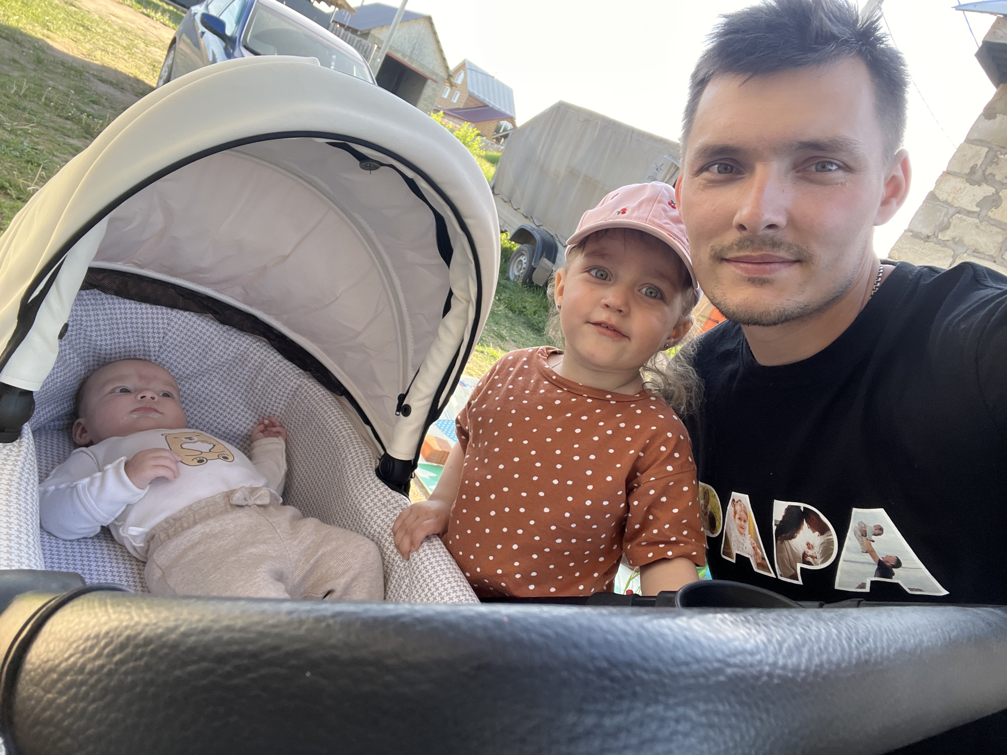 The image shows a man taking a selfie with two young children. One child is a baby lying in a white stroller with a patterned grey and white liner. The baby is wearing a white long-sleeved shirt with a bear on it and beige pants. The second child, a toddler, stands next to the stroller wearing a pink baseball cap and a brown polka-dot shirt. The man is wearing a black t-shirt with the word "PAPA" designed with pictures in each letter.
The background includes a grassy area, a building with a gray metal roof, and a parked vehicle that looks like a trailer. The location is Chaikovsky, Russia.