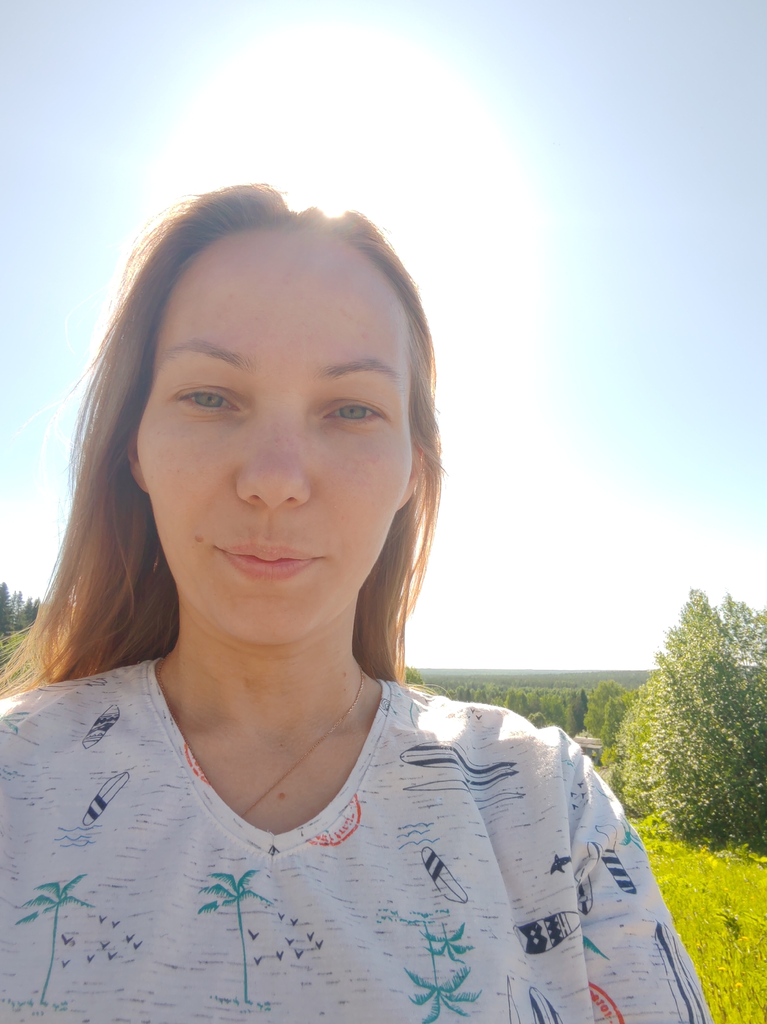 A female subject is depicted in a close-up outdoor selfie, facing the camera with a neutral expression. She has long, light brown hair, green eyes, and fair skin, with a visible mole on her left cheek. She wears a light grey V-neck t-shirt patterned with blue and green illustrations of palm trees, surfboards, and small birds. A thin gold chain necklace is visible around her neck. The background consists of a clear, bright blue sky with a strong sun glare originating from the upper left, causing a lens flare. Beyond the subject, a distant line of dense green forest extends across the horizon. To the right, a brightly sunlit deciduous tree and yellow wildflowers in green grass are visible. The scene is located in Чухлэм, Russia.