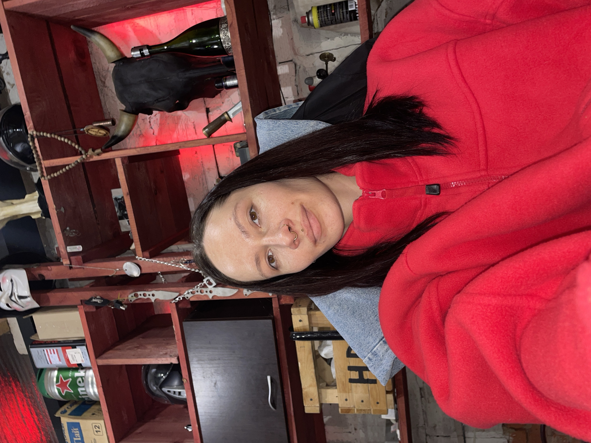 The image shows a person wearing a red fleece jacket and a light blue denim jacket. The person has dark hair and facial piercings (nose and lip). Behind the person is a red wooden shelf unit containing various items, including a bull skull with attached wine bottle, a helmet, boxes of beer, and knives. The background suggests a room with concrete walls. There is no evidence to confirm the location.