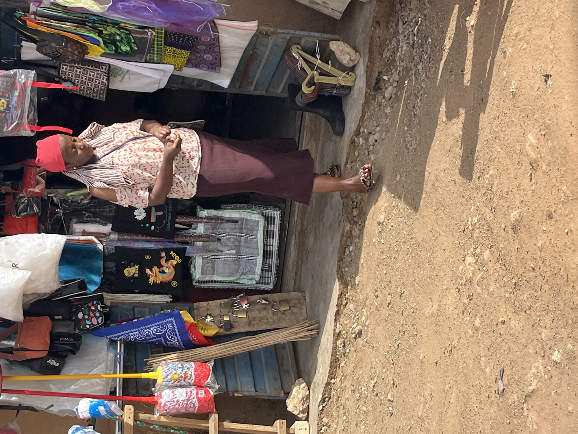 The image shows a woman standing in what appears to be a small shop. The shop is filled with various items including bags, purses, mops, and clothing. The woman has on a red head covering and is wearing sandals. Outside the shop is a dirt area with a long shadow cast onto it. This shop is located in Jos, Nigeria.