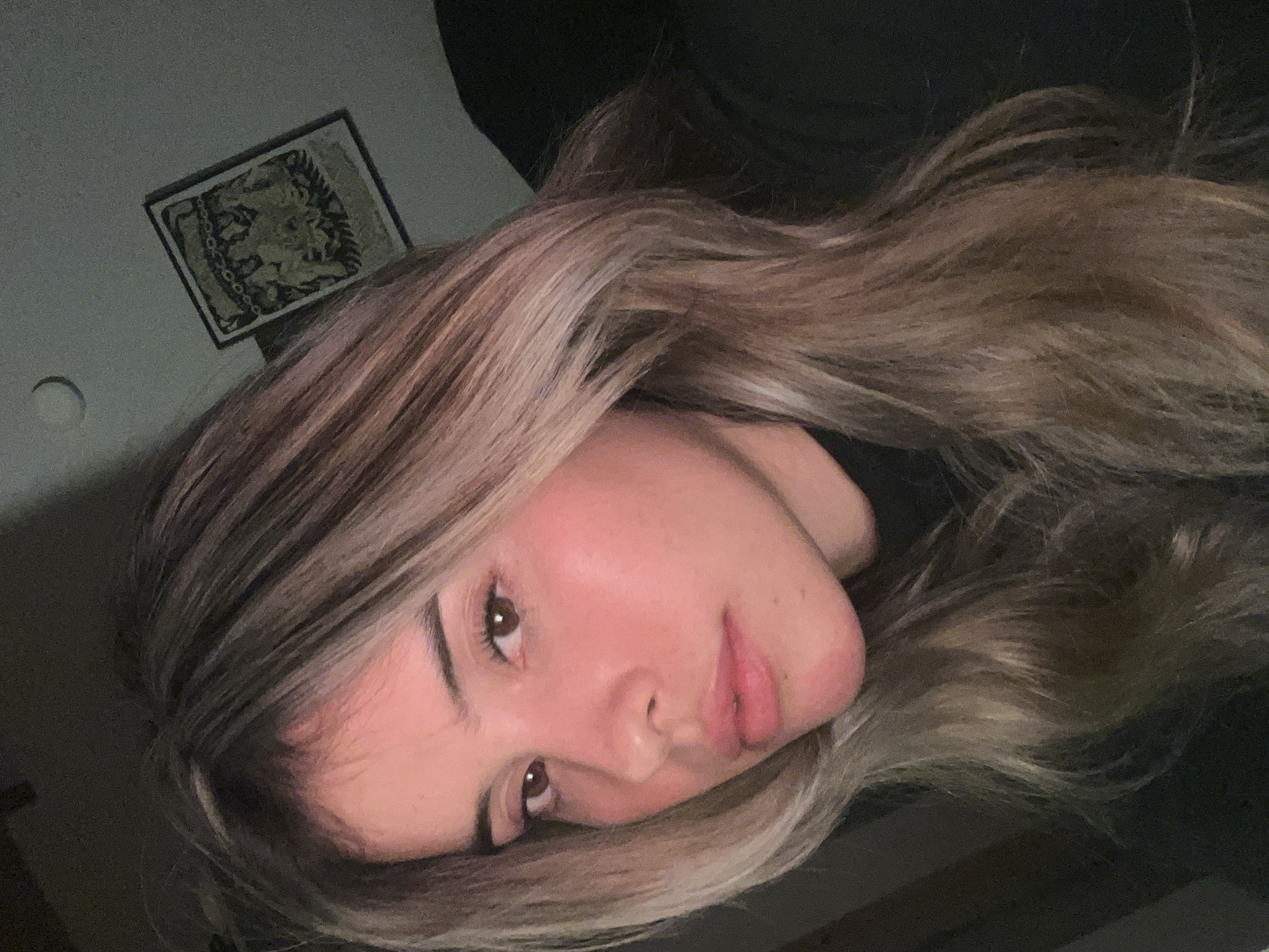 A woman is posing for a selfie indoors. The background includes a dark wall with a framed artwork hanging on it, and a circular light fixture on the ceiling. The woman has long, light brown hair with blonde highlights, and is wearing a black garment. Her facial features are visible, with her head tilted slightly to the side. The lighting is warm and appears to be artificial. There is no indication of the location being in Sumalao, Argentina.