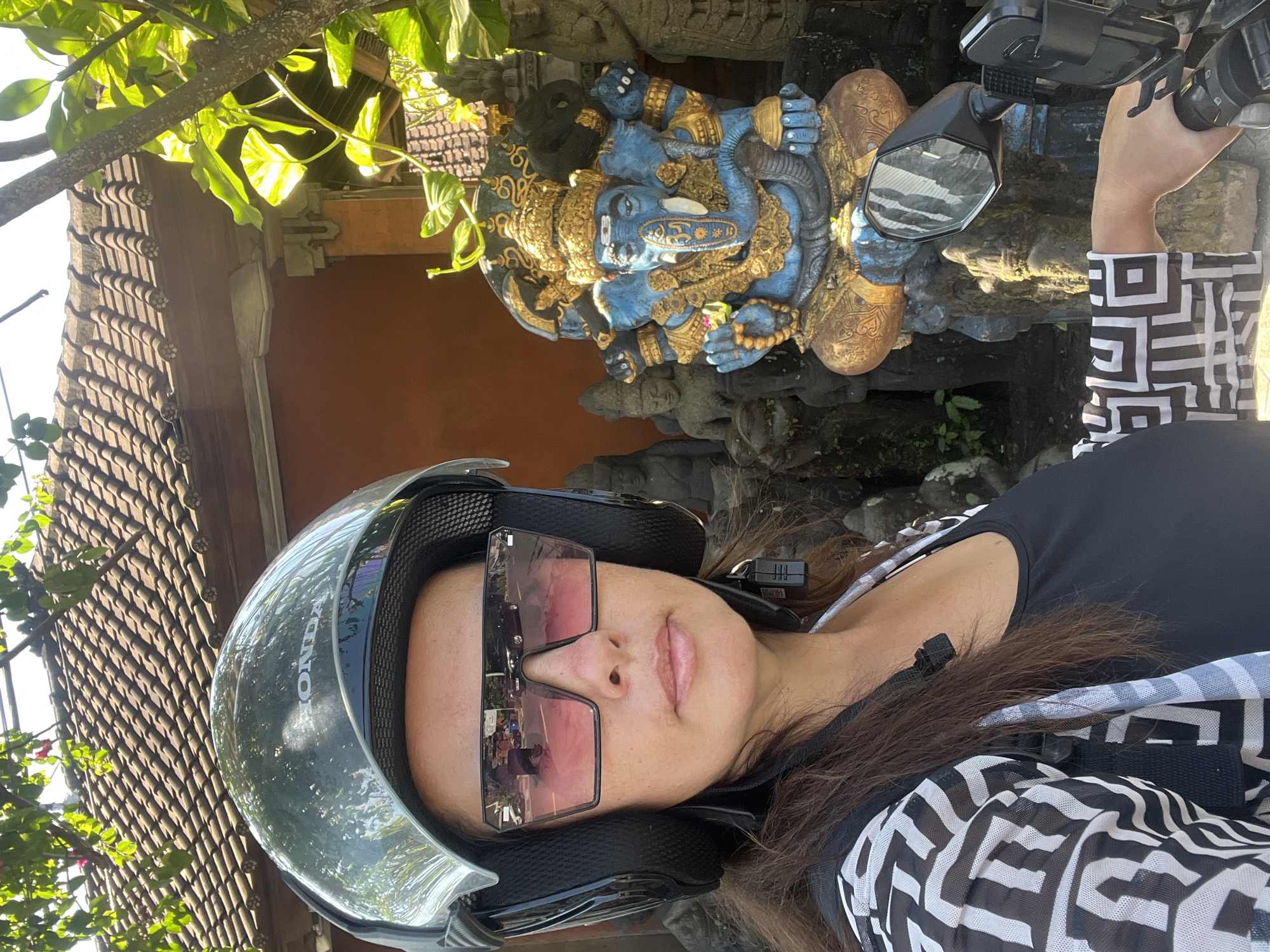 A woman wearing a helmet and large sunglasses is taking a selfie on a scooter. She is dressed in a black tank top and a black and white patterned shirt. Behind her, to the right, is a statue of Ganesha, a blue elephant-headed deity. The statue is adorned with gold jewelry and intricate detailing. Several other stone statues are visible in the background, along with a building with a tiled roof and trees. The scene appears to be outdoors, possibly in the city of Sukawati, Indonesia, given the cultural context of the statues.
