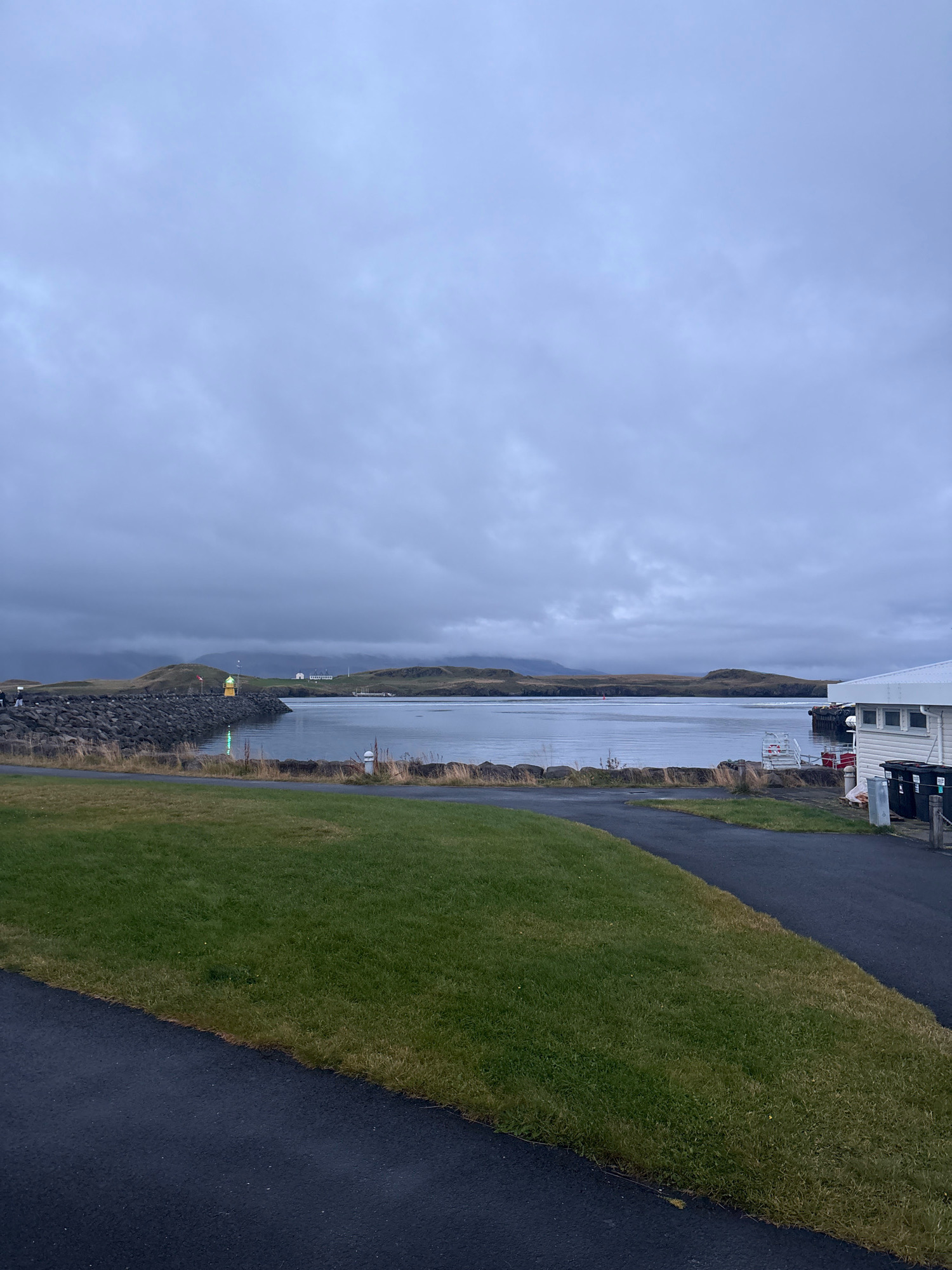 The image depicts a somber, overcast scene at a harbor, likely in or near Reykjavik, Iceland, given the description of the location. A breakwater made of dark, irregular rocks extends into a calm body of water, with a lighthouse visible on its far end. The water is a dull gray, reflecting the cloudy sky. Across the water, low, rolling hills form the backdrop, with a few scattered white buildings suggesting a small settlement or coastal infrastructure.
In the foreground, a paved pathway curves through a well-maintained patch of green grass. To the right, a white building with a dark roof and windows stands adjacent to a set of black trash bins. The overall atmosphere is one of quiet solitude and natural beauty, characteristic of a coastal Icelandic landscape. No people are clearly visible in the frame.