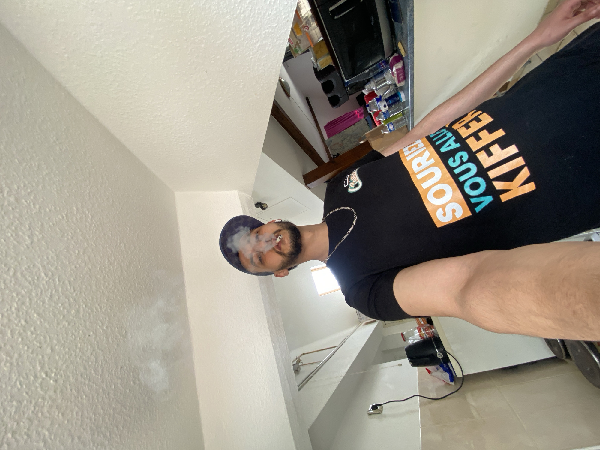 A man with a beard and wearing a dark baseball cap and a black t-shirt that reads "Sourire Vous Allez Kiffer" is smoking, with smoke exhaling from his mouth. He is also wearing a silver chain. The photo appears to be taken in an interior location with plain white walls. The background includes a glimpse into what looks like a kitchen or pantry area with shelves, appliances, and supplies. A staircase is visible in the background. There is no information to indicate that the photo was taken in Bischheim, France.