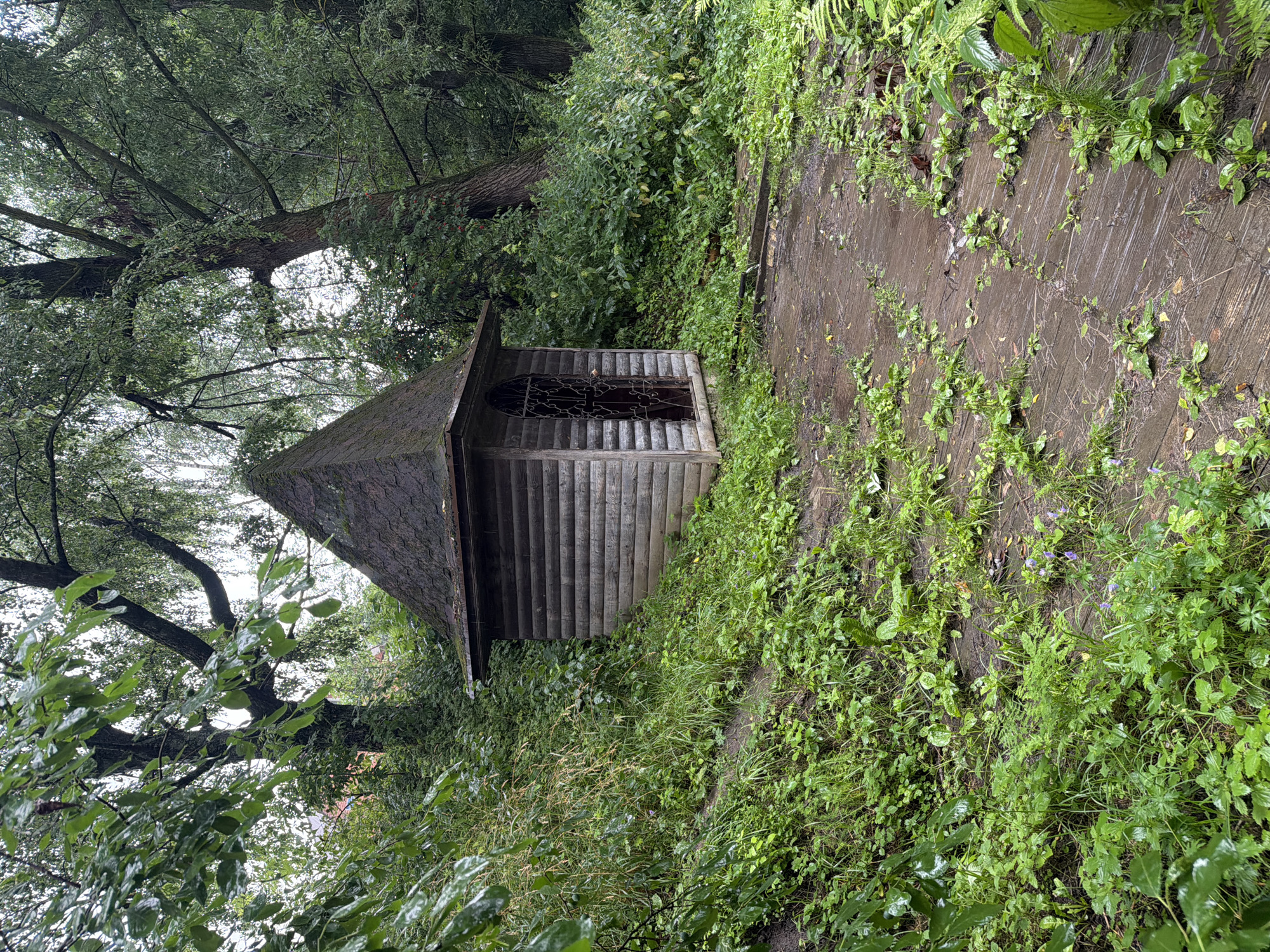 In Nikulskoye, Russia, a small, weathered wooden structure with a pyramidal roof stands amidst lush greenery. The building, constructed from horizontal wooden planks, features a single entrance with an ornate metal grate. The surrounding area is a dense forest with a path made of wooden planks that appears damp and overgrown with grass and small plants. The trees are tall and their branches, laden with leaves, create a canopy overhead, suggesting a secluded, natural setting. The scene is peaceful and appears to be after a rainfall, as indicated by the wetness of the ground and foliage. There are no people or animals visible in the image.