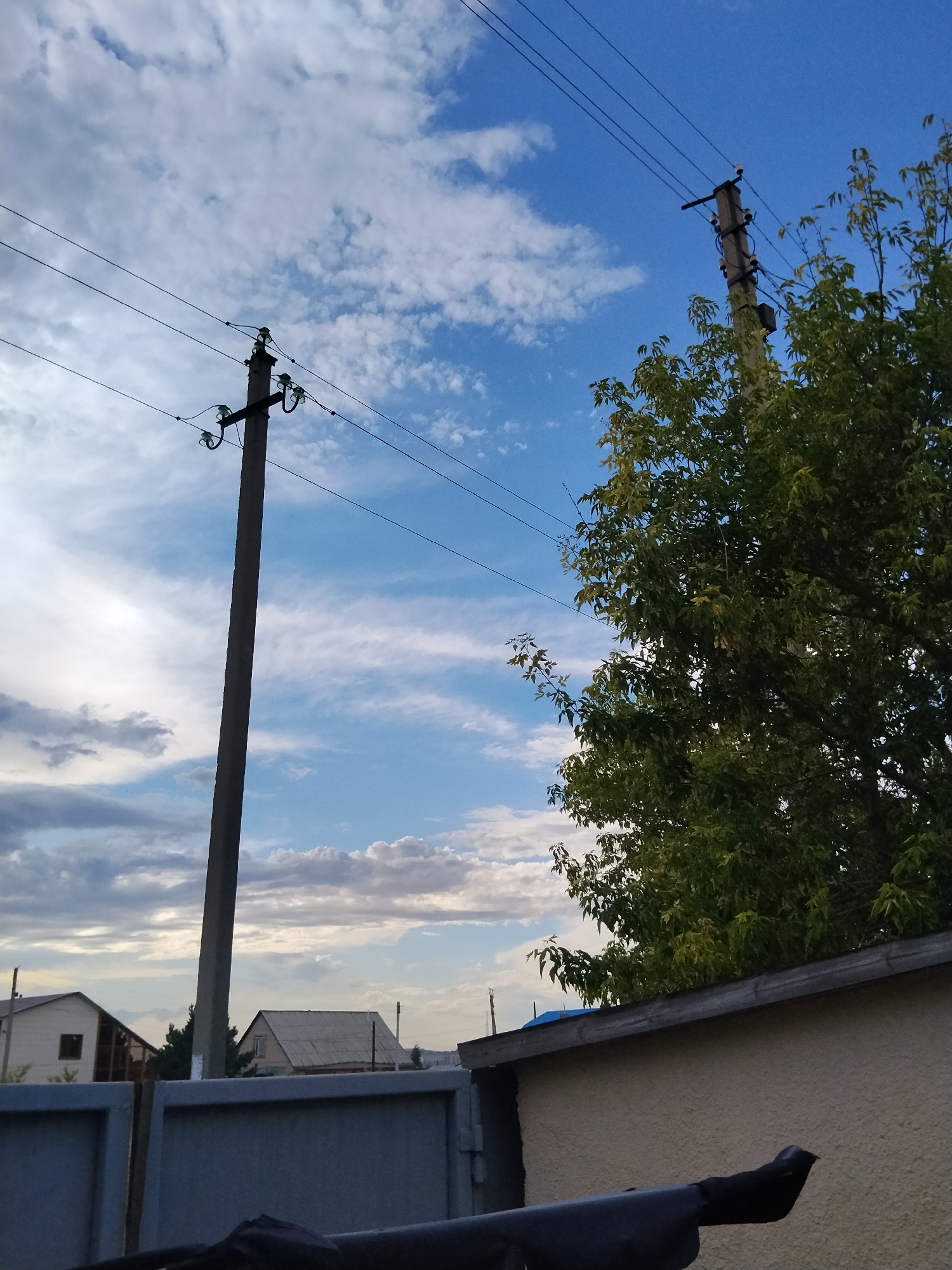The image shows a daytime exterior scene with a bright blue sky filled with scattered white clouds. In the foreground, a dark gray metal fence is visible, with a black object draped over its top. Behind the fence, a textured light-colored wall of a building runs across the lower right of the frame. To the left of the building, a tall, slender concrete utility pole stands prominently, supporting several black power lines that stretch across the sky. Another similar utility pole is visible further in the background to the right, partially obscured by the dense green foliage of a large tree. The tree, with its vibrant green leaves, occupies the upper right quadrant of the image, its branches reaching towards the sky. In the background, beyond the fence and utility poles, two residential houses are visible. The house on the left is light-colored with a dark-roofed section, and the house on the right has a grey, corrugated roof. The overall scene suggests a suburban or semi-rural environment. The specific location is identified as being in Kokshetau, Kazakhstan.