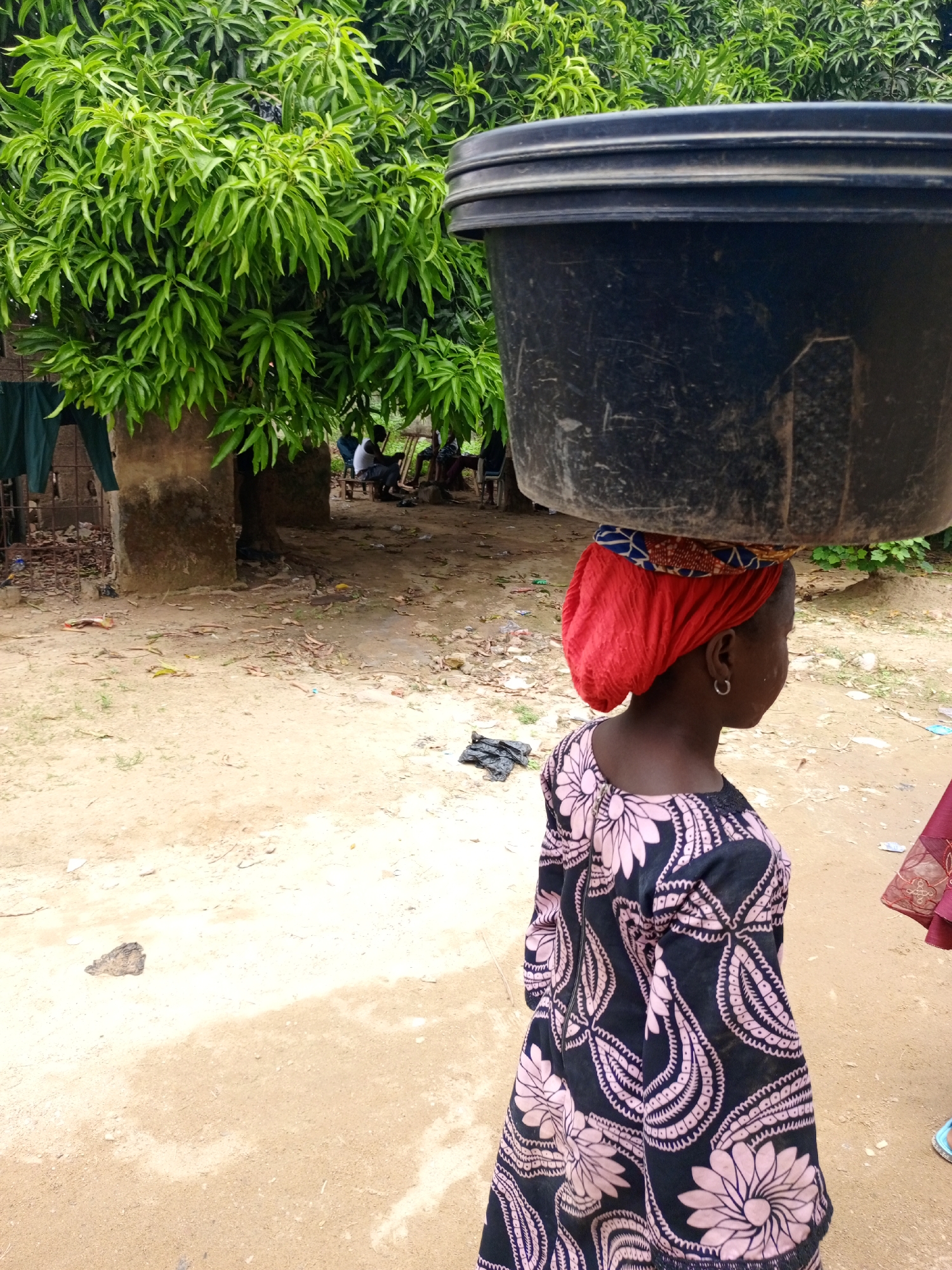The image depicts a young girl in Keffi, Nigeria, carrying a large black bucket on her head. She is wearing a red headscarf and a black dress with a pink floral pattern. The background includes a dirt road, a tree with lush green foliage, and a partially visible building. In the distance, there are people sitting outside in an open space. The environment suggests a rural or less developed area within the city.