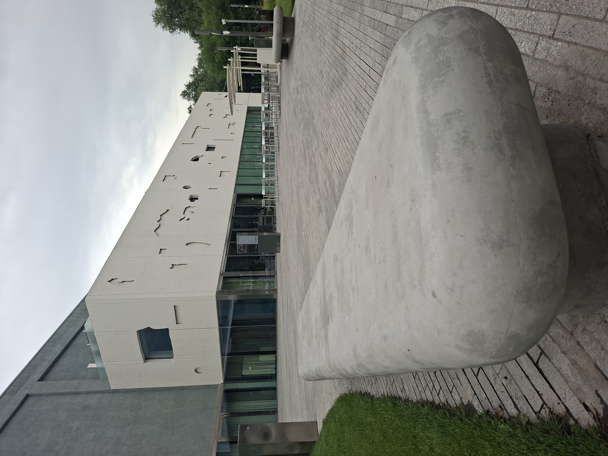 An exterior shot shows a modern building with a gray textured facade and a white facade with irregular cutouts and a single rectangular window. The white facade runs horizontally along the ground floor and features large glass windows. In front of the building is a paved outdoor area with a long, smooth concrete bench in the foreground. To the left of the bench is a strip of green grass. In the background, there are trees and an overcast sky. The location is identified as the Central State Museum of Kazakhstan in Almaty, Kazakhstan. There are no people visible in the image.