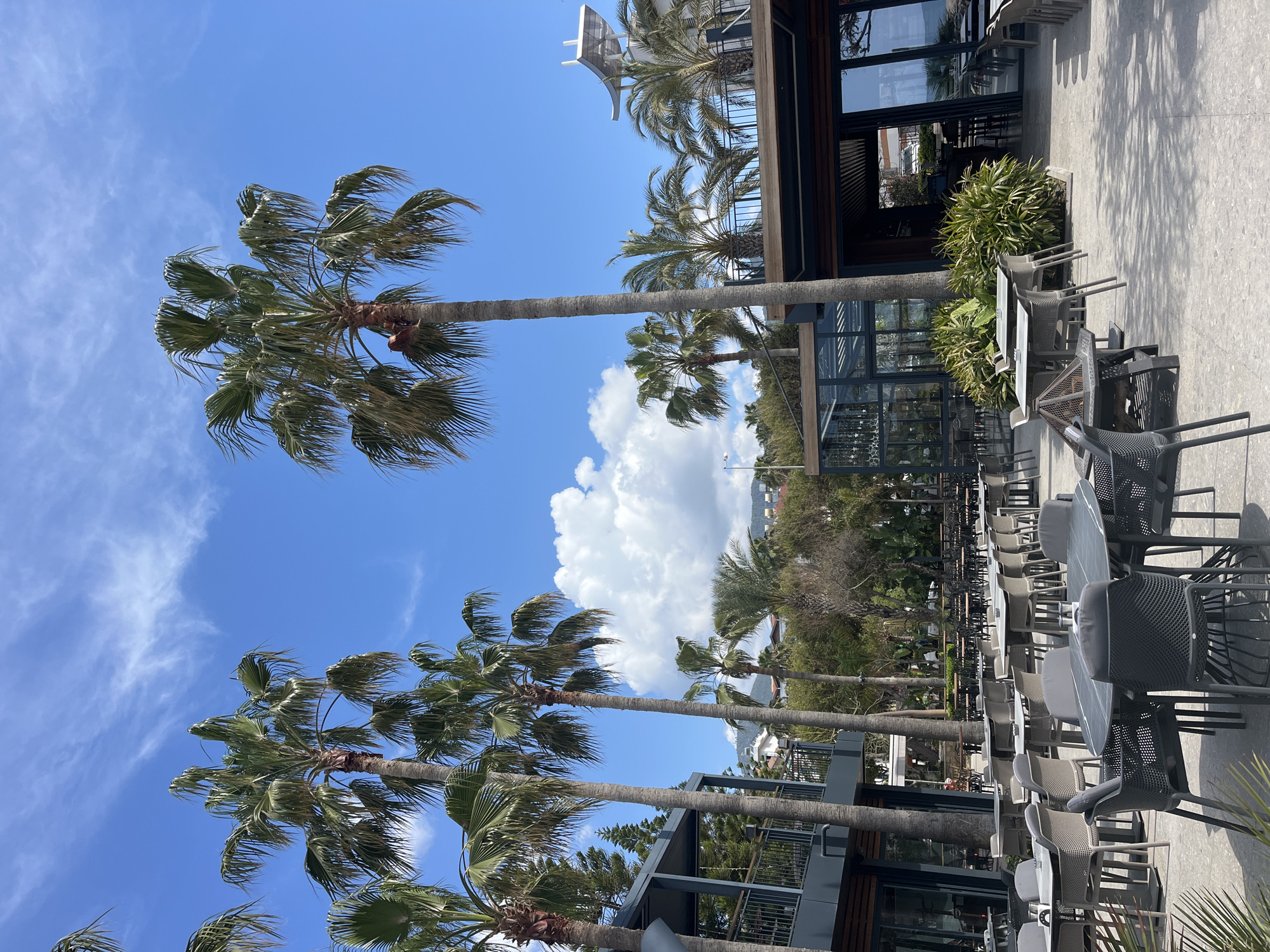 The image shows an outdoor dining area with numerous tables and chairs, located in Alanya, Turkey, as palm trees are visible in the background. The location seems to be adjacent to a modern building with glass windows and an upper level, possibly a restaurant or cafe. The sky is blue with scattered clouds, and the sun is shining, casting shadows on the ground. There are no people visible in the immediate foreground.