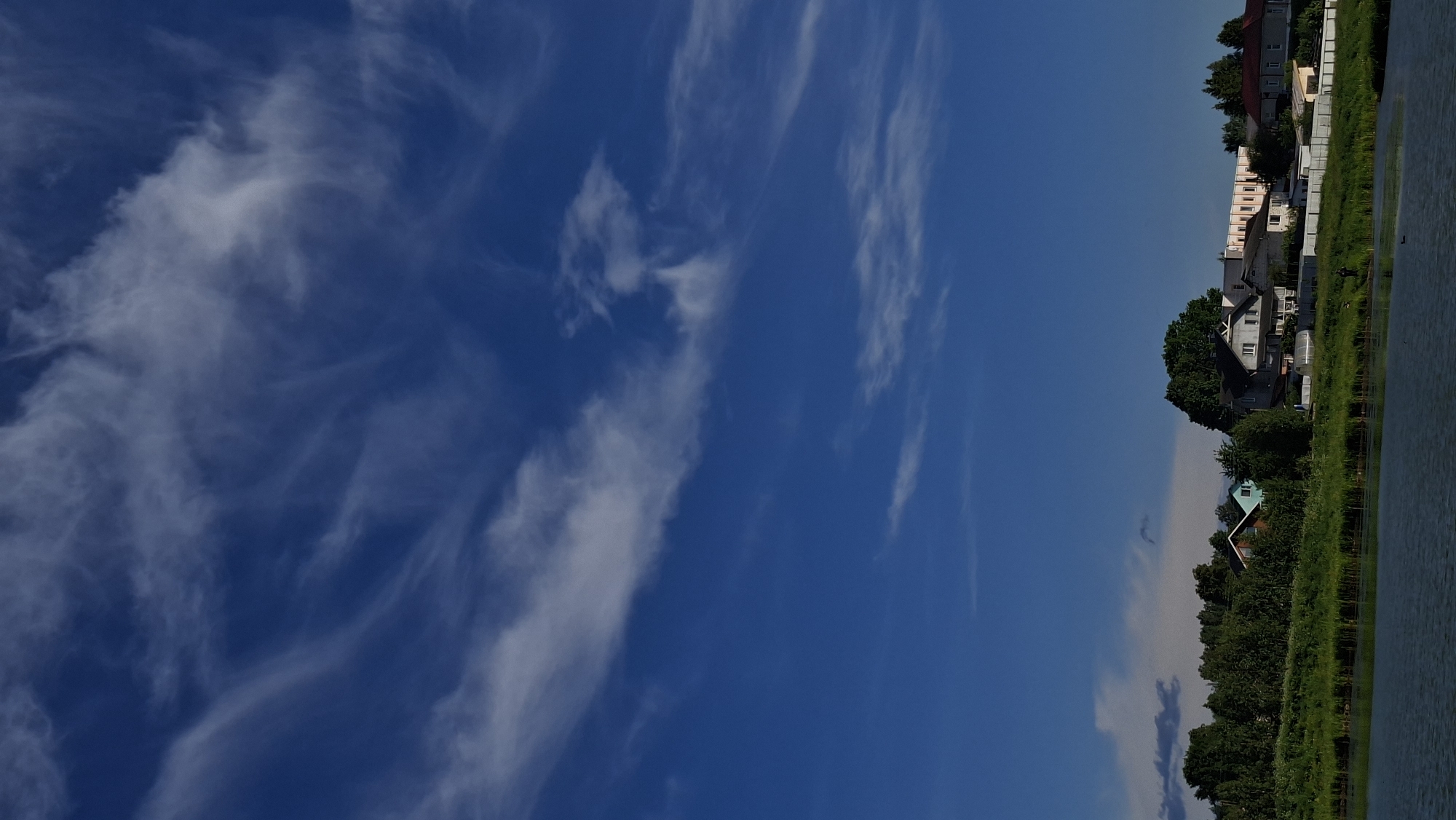 The image captures a peaceful daytime landscape, likely near Забалоцкі сельскі Савет, Belarus, under a vast blue sky. The upper two-thirds of the frame are dominated by a clear, vibrant blue sky, featuring scattered, wispy white clouds that stretch and swirl, indicative of cirrus or cirrocumulus formations.
The lower third of the image showcases a tranquil body of water, possibly a lake or river, on the right side. A lush band of green vegetation, composed of trees, tall grasses, and reeds, lines the water's edge, creating a natural boundary. Behind this greenery, a collection of residential buildings, primarily houses, stretches across the landscape. These structures vary in appearance; some have light-colored facades, while others are darker. Notable details include a house with a distinctive greenish roof in the foreground, and other houses with dark roofs. A fence or wall appears to run alongside some of the properties.
The scene suggests a bright, sunny day, with no visible signs of rain or heavy cloud cover. The strong daylight illuminates the landscape clearly. There are no people or specific activities depicted, contributing to the serene and calm atmosphere of this natural and semi-rural setting. No text is visible in the image.