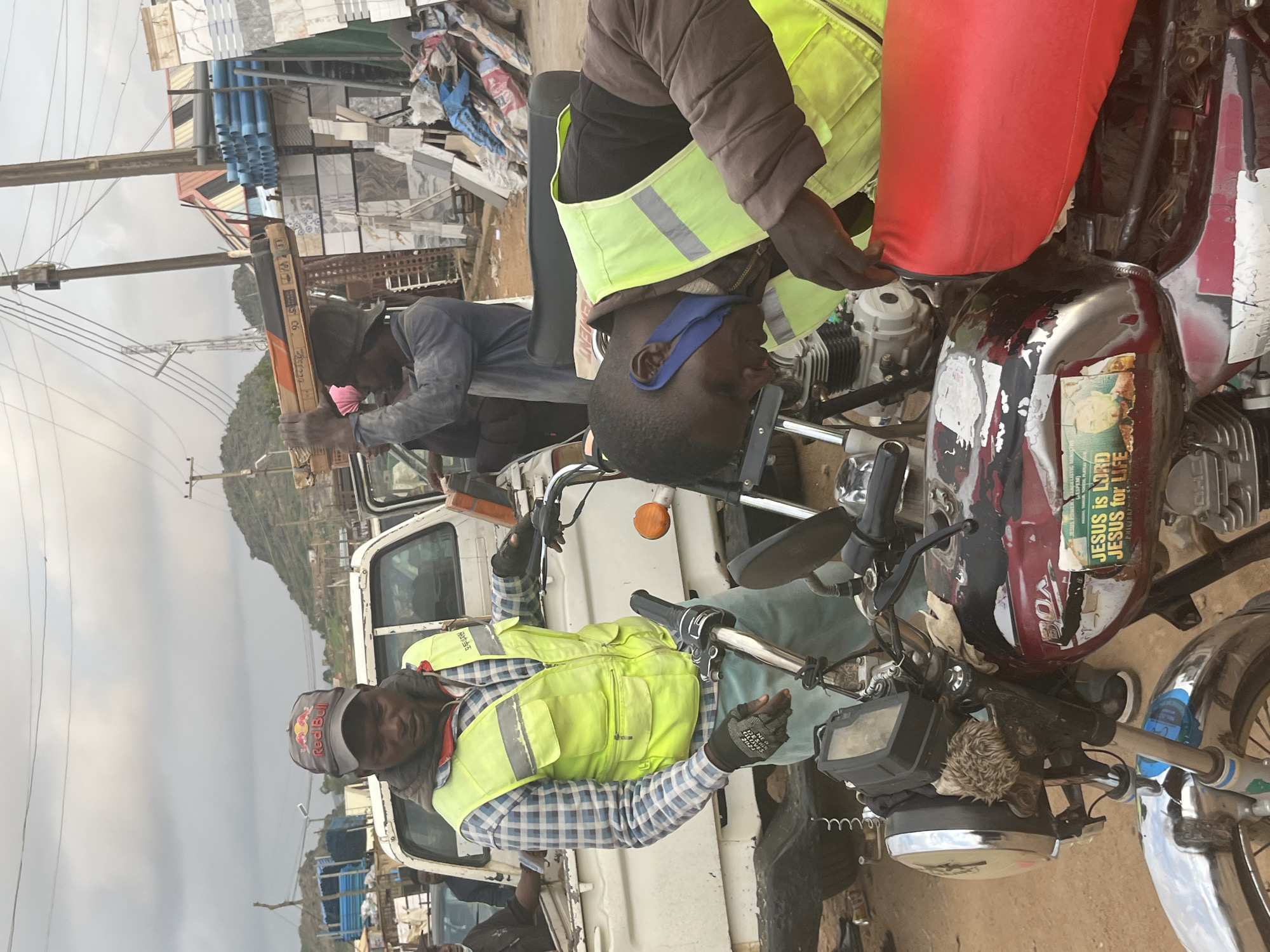 The image captures an outdoor scene in Gura Top, Nigeria, where two men are working on a motorcycle. The man on the left, wearing a Red Bull cap, a plaid shirt, and a high-visibility vest, is interacting with the motorcycle's handlebars. His right hand is gloved and positioned near the front brake lever. He appears to be instructing or assisting the second man.
The second man, on the right, is leaning over the motorcycle, focusing on its engine area. He is wearing a high-visibility vest over a brown jacket and has a blue cloth tied around his ear. His hands are engaged with the motorcycle's components, suggesting he is performing a repair or maintenance task.
The motorcycle itself is red and black, showing signs of wear and tear with peeling paint and various stickers. One prominent sticker features the text "JESUS is LORD JESUS for LIFE."
The background reveals a bustling environment with multiple vehicles, including a white truck and a blue truck laden with pipes. Stacks of tiles and other building materials are visible, along with utility poles and wires. The terrain appears to be unpaved, indicative of a roadside or market area. The sky is overcast. In the distance, a hilly landscape is visible.