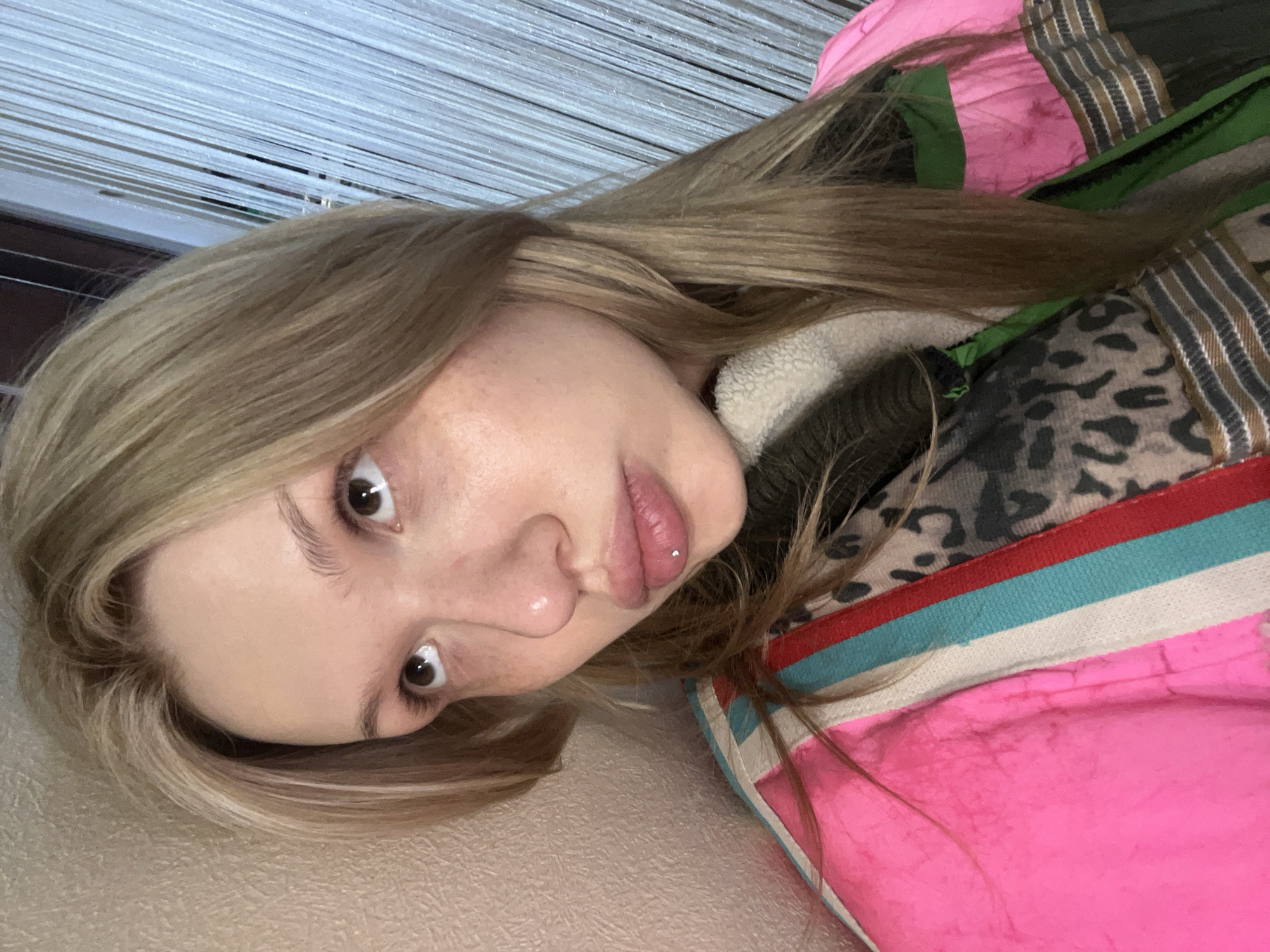 A young individual, presumed female, with fair skin and long light brown hair, is depicted in a close-up, head-and-shoulders shot against an indoor background in Chaikovsky, Russia. The subject's dark brown eyes are directed upwards and slightly to the right of the frame. A silver stud labret piercing is centered on the lower lip. The individual is wearing a multi-layered jacket. The dominant outer layer is a neon pink fabric, textured with wrinkles, featuring a vertical stripe pattern on the left side composed of white, light blue, and red bands. Beneath this, a dark olive or green garment with a white, sherpa-like collar is visible on the right side of the neck and shoulder. Further to the right, a section of fabric with a dark grey and beige leopard print is visible, alongside a dark-colored garment featuring horizontal stripes in brown, beige, and grey tones, and a green zipper or trim. The background on the left consists of a textured, light beige wall. On the right, a translucent curtain composed of numerous thin, white vertical strands is present.