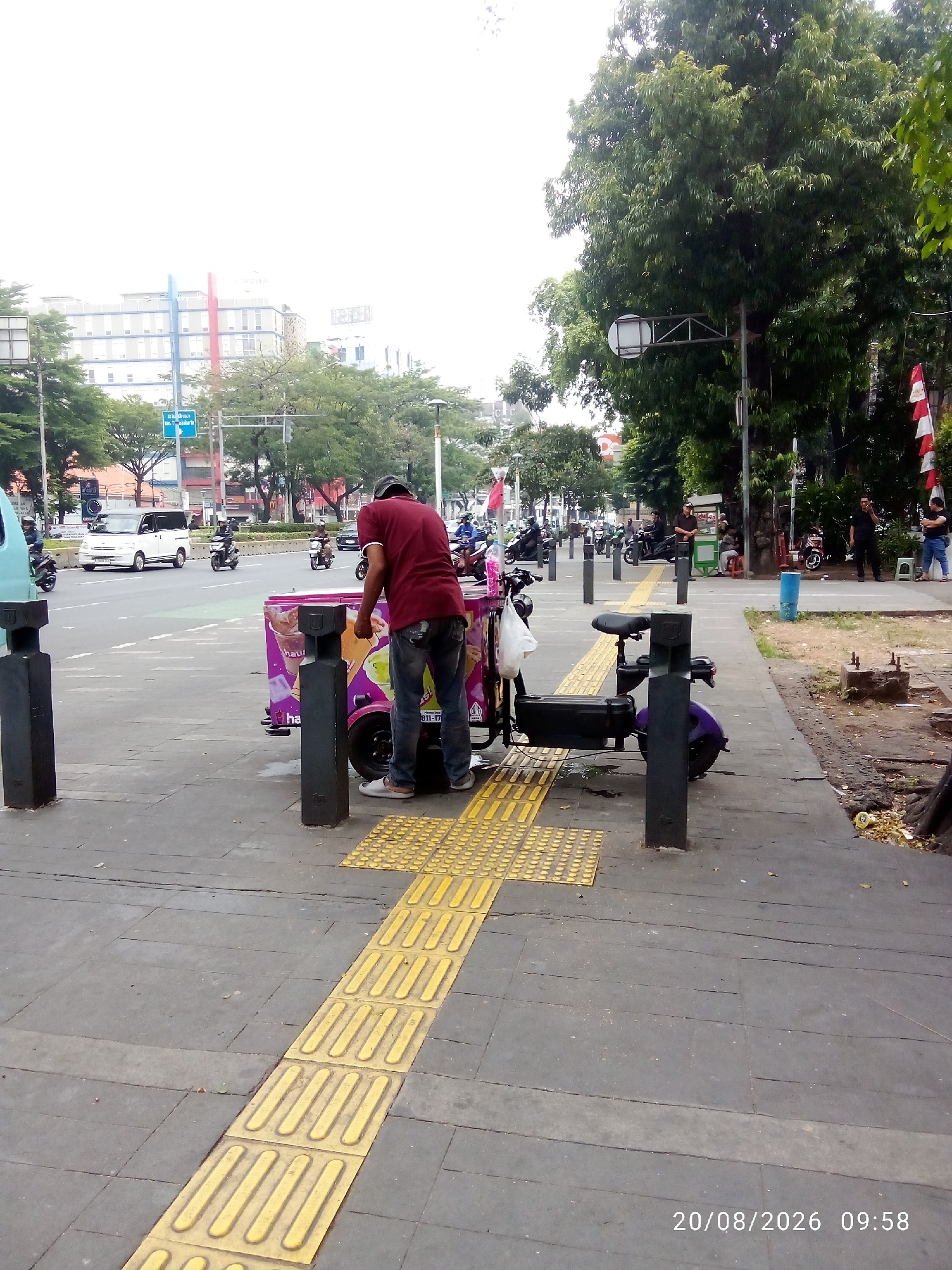 A man wearing a maroon t-shirt and blue jeans stands next to a purple and pink ice cream cart on a paved sidewalk. He is bent over, appearing to be working with the cart. A purple electric scooter is parked beside the cart. The sidewalk is bordered by tactile paving for the visually impaired. In the background, a street with traffic, including cars and motorcycles, is visible. Trees and buildings line the street. A street light is also present. The image is timestamped "20/08/2026 09:58".