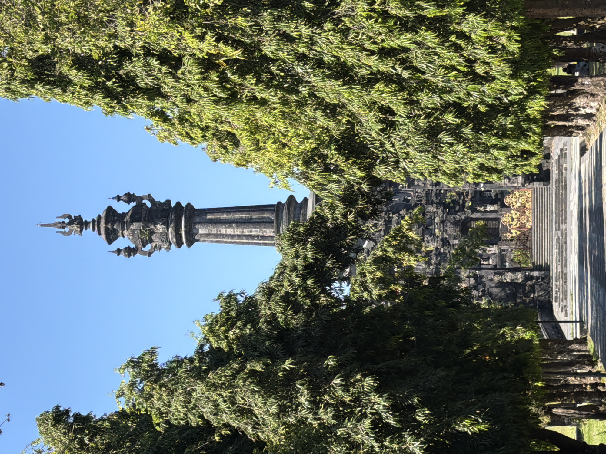 This image depicts a tall, ornate stone tower, possibly a temple or monument, set against a clear blue sky. Lush green trees frame the tower, with some branches extending into the foreground and others partially obscuring the structure. The architecture of the tower is intricate, featuring multiple tiers and decorative elements, including spires and what appear to be carved figures or ornaments.
To the right of the tower, a pathway or staircase with stone steps is visible, leading away from the viewer and into the shadows of surrounding structures. The stonework of these buildings appears weathered and ancient, suggesting a historical or cultural site. The presence of sunlight filtering through the leaves and casting shadows indicates it is daytime, likely with fair weather given the clear sky.
In the background, on the right side of the image, two figures can be seen walking along the paved area, adding a sense of scale and human presence to the scene. The overall atmosphere is serene and perhaps a little awe-inspiring, due to the grandeur of the architecture and the natural beauty of the foliage. There is no visible text in the image. The location context is Denpasar, Indonesia, which aligns with the architectural style and tropical vegetation.