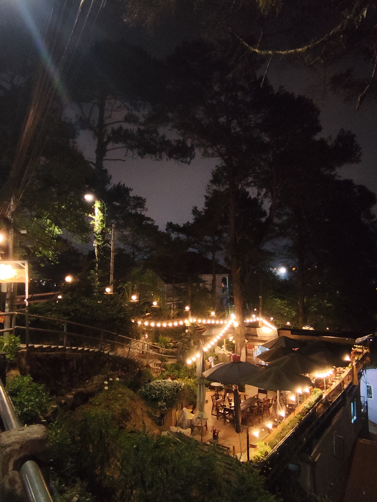 A nighttime shot showcases a cafe or restaurant setting in what appears to be Da Lat, Vietnam. The camera's vantage point is elevated, looking down at the terraced outdoor seating. String lights illuminate the scene, adding a warm glow to the tables and the surrounding greenery. Several dark umbrellas are positioned over tables, suggesting patrons are seated underneath. The architecture is typical of mountainous regions, with stepped stone pathways and building construction clinging to the hillside. Tall pine trees frame the scene, silhouetted against the night sky. Streetlights are visible in the background, marking the landscape. No individuals are clearly visible, making it difficult to assess activity. The focus remains on the ambiance of the venue and its integration with the natural landscape.
