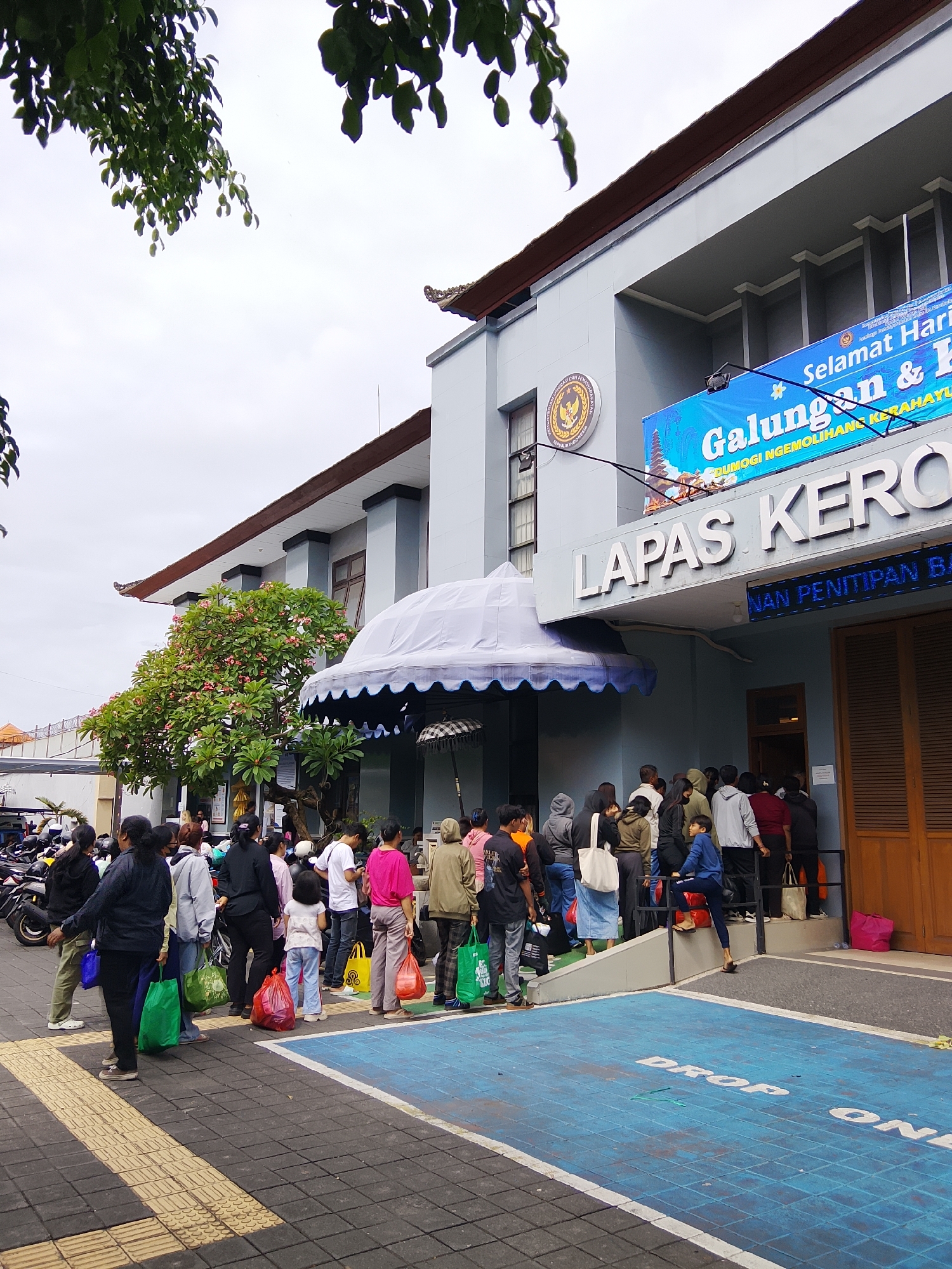 The image depicts the exterior of a government building, likely a correctional facility, in North Kuta, Indonesia. A queue of approximately 20-25 individuals, varying in age and gender, stands outside the building. They are dressed in casual attire and many are carrying bags, some of which appear to be grocery bags or bags containing personal belongings. The building itself is a multi-story structure with a light blue facade and a dark brown roof. A prominent banner is displayed across the building, bearing text in Indonesian and an image of a traditional Balinese landscape. The words "LAPAS KERO" are visible in large white lettering on the facade. The ground in front of the building is paved with dark gray and yellow tiles, with a blue section marked with white text reading "DROP ON". A tree with pink flowers is visible to the left of the entrance, and several motorcycles are parked nearby. The sky is overcast.
