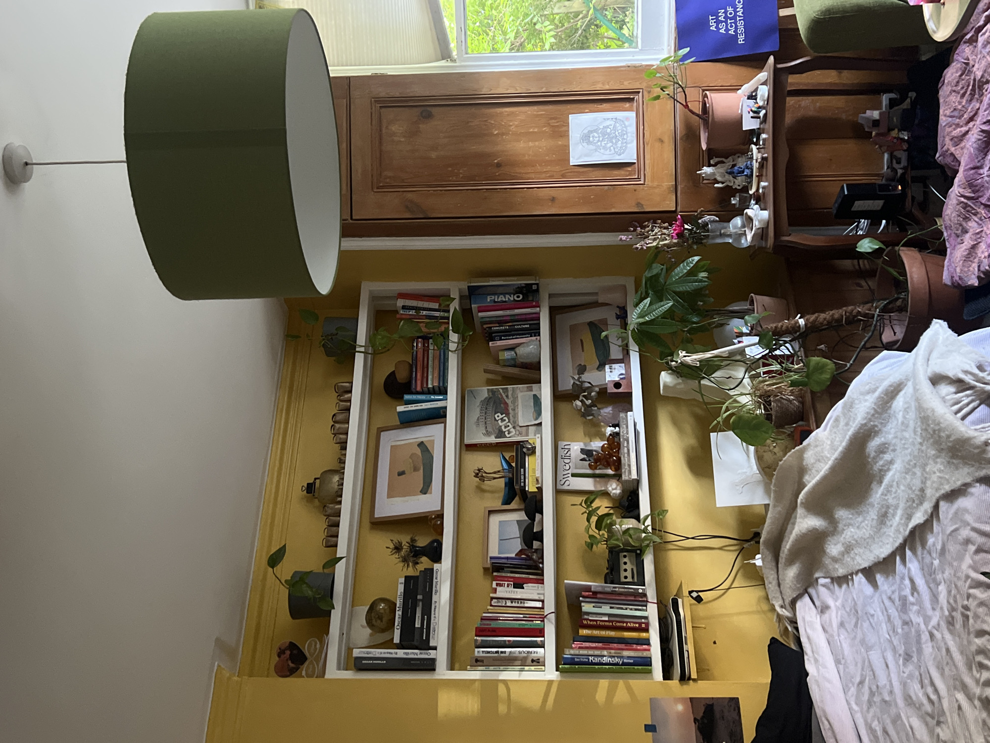 The image depicts an interior scene, possibly a bedroom, with yellow walls and a large, olive-green lampshade hanging from the ceiling. A white bookshelf unit is filled with books, plants, and decorative items. To the right, a wooden cabinet stands next to a window with venetian blinds, through which green foliage is visible. A small wooden table next to the cabinet holds various objects, including figurines and candles. A bed with a patterned duvet and a fluffy throw is partially visible in the foreground on the left. A blue poster with white text that reads "ART AS AN ACT OF RESISTANCE" is leaning against the wall to the right of the cabinet. The scene is well-lit, with natural light coming from the window and overhead lighting. The overall impression is one of a lived-in, eclectic space. There is no visible indication of the specific location within London, United Kingdom.